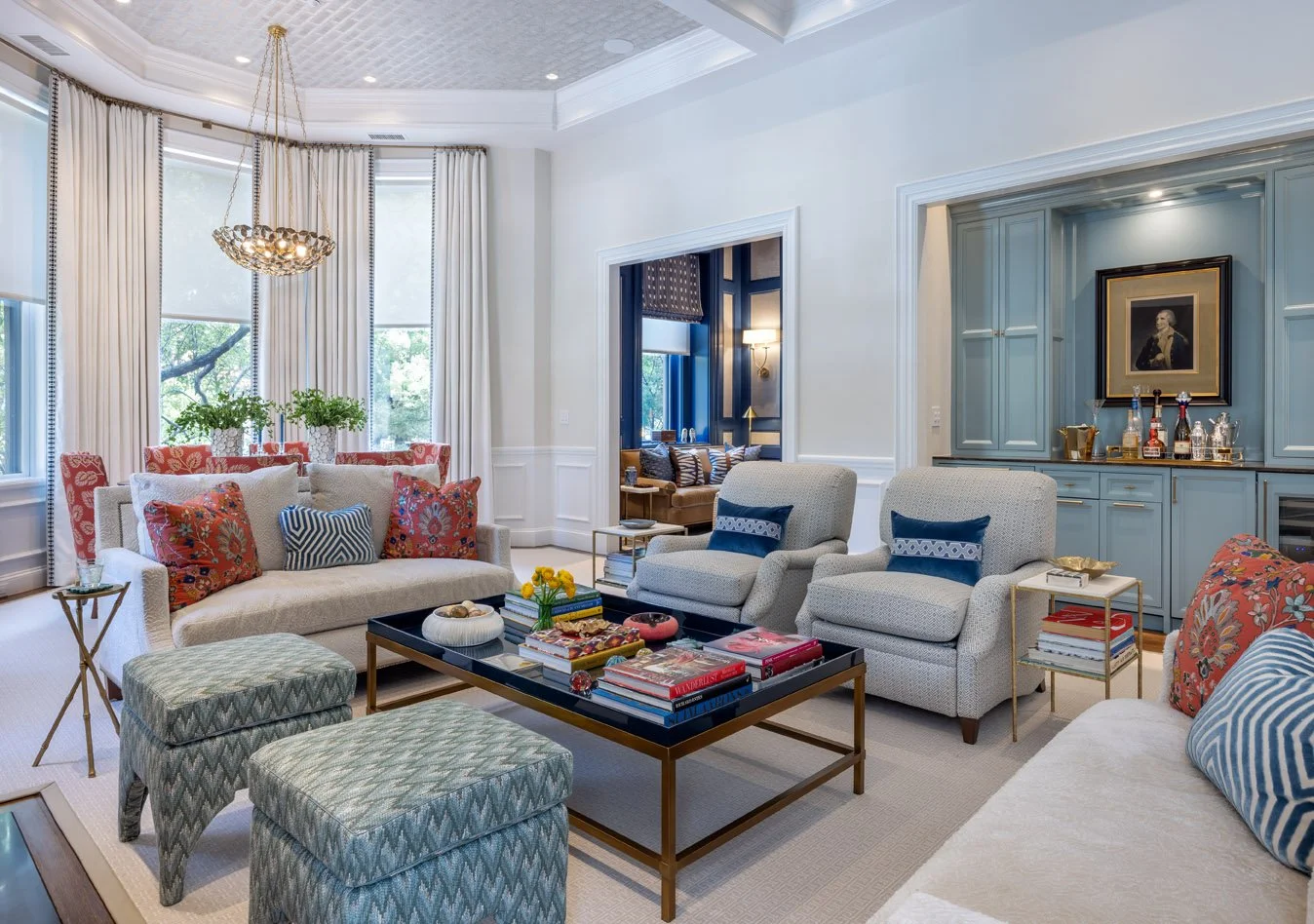A spacious living room with white walls, large windows with white curtains, and elegant furniture. There are multiple sofas and armchairs with decorative pillows, a black coffee table with books and decorative items, and a blue built-in bar area with framed artwork. The room features a high ceiling with a chandelier and a view of greenery outside.