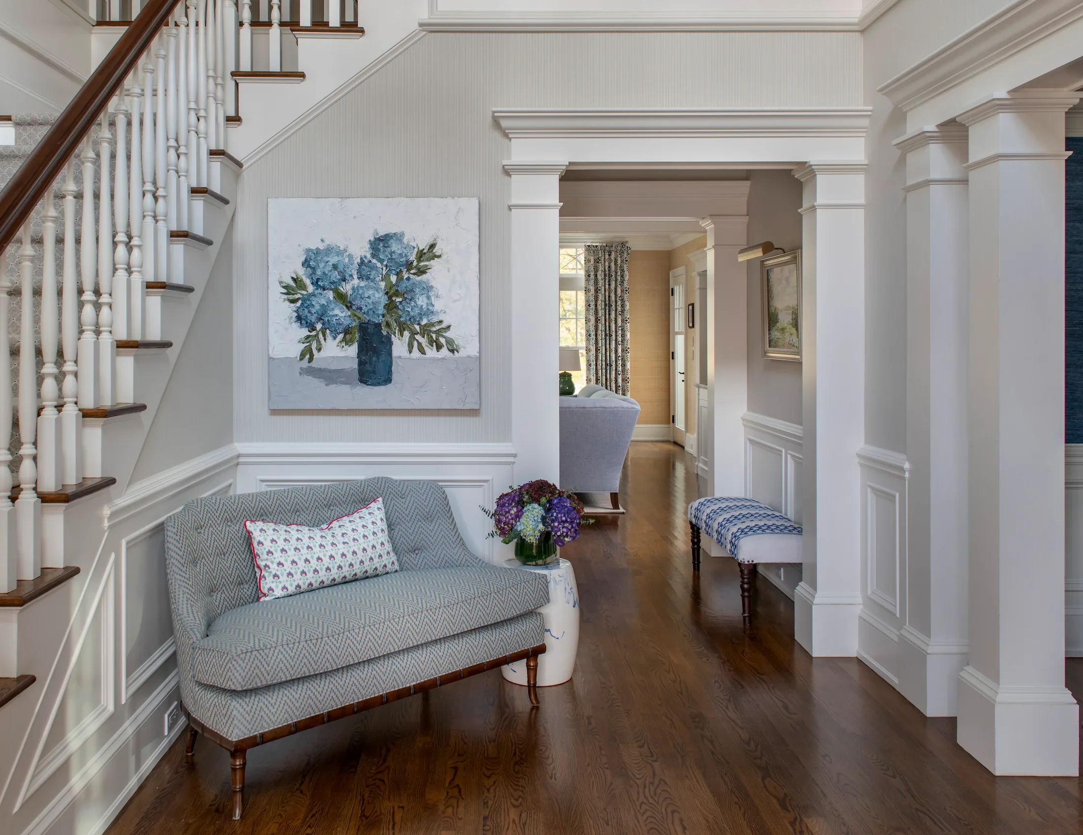 A living room corner with a plaid-patterned sofa, a glass-topped side table with a vase of purple flowers, and a large painting of blue flowers in a dark vase hanging on the wall. There is a staircase on the left and a doorway leading to another room with a window and patterned curtains.