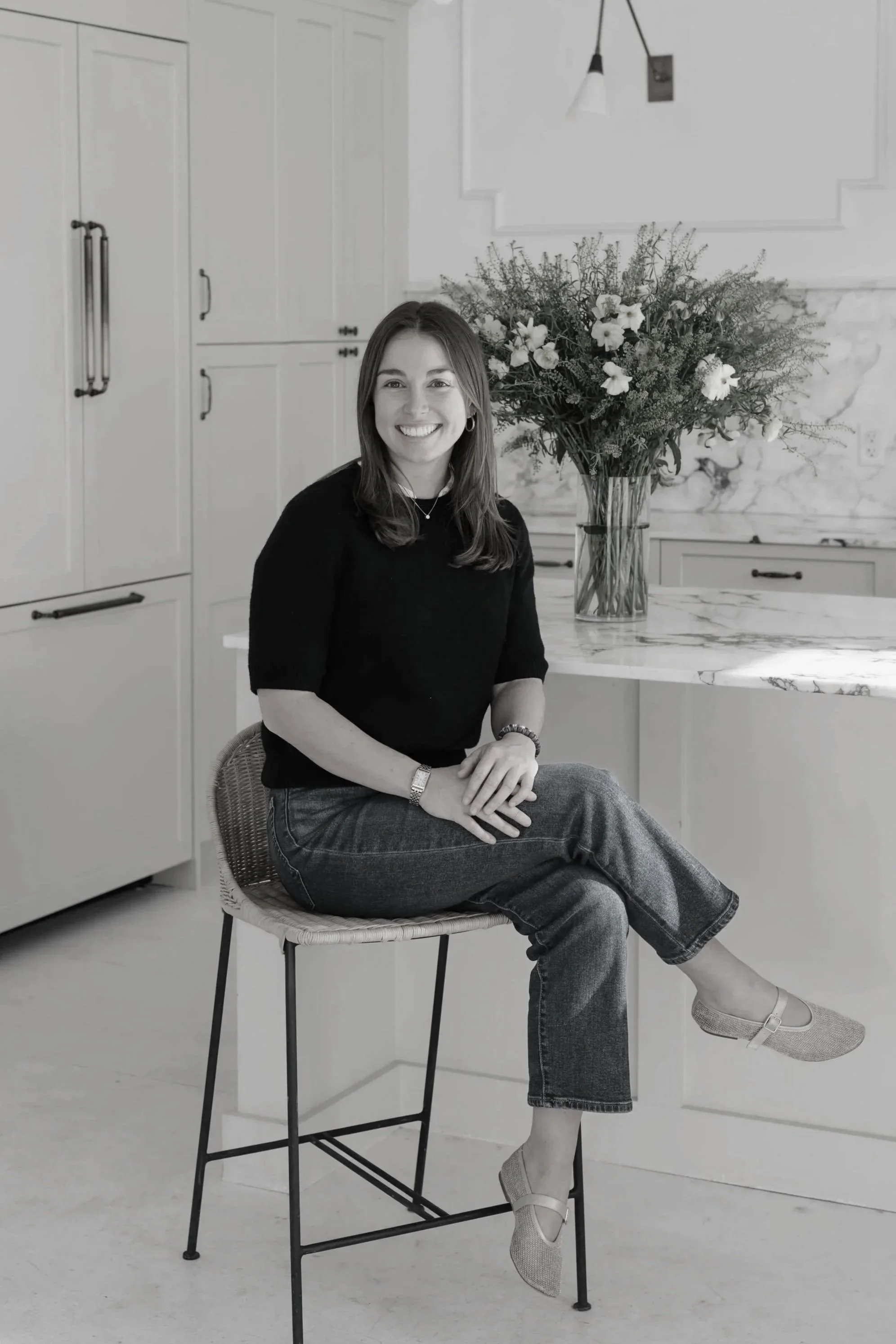 A young woman with shoulder-length hair and earrings sitting on a bar stool in a bright, modern kitchen with white cabinets, a marble kitchen island, a large bouquet of flowers in a glass vase on the counter, and a modern light fixture hanging from the ceiling. She is smiling, wearing a black top, jeans, and flat shoes.