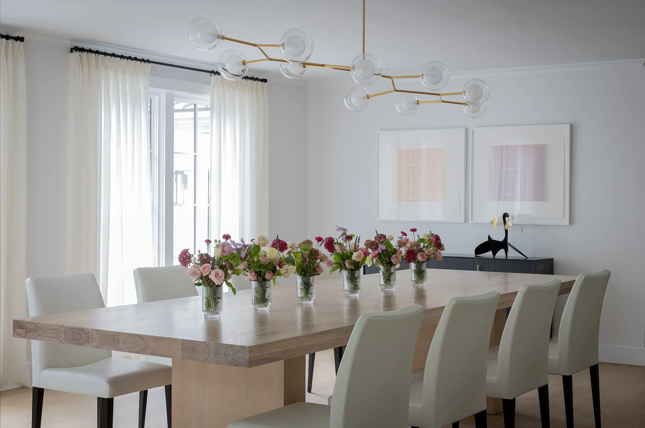 A modern dining room with a large wooden table, white chairs, and multiple glass vases filled with pink, purple, and red flowers. A contemporary chandelier hangs above, and there are white curtains covering large windows in the background.