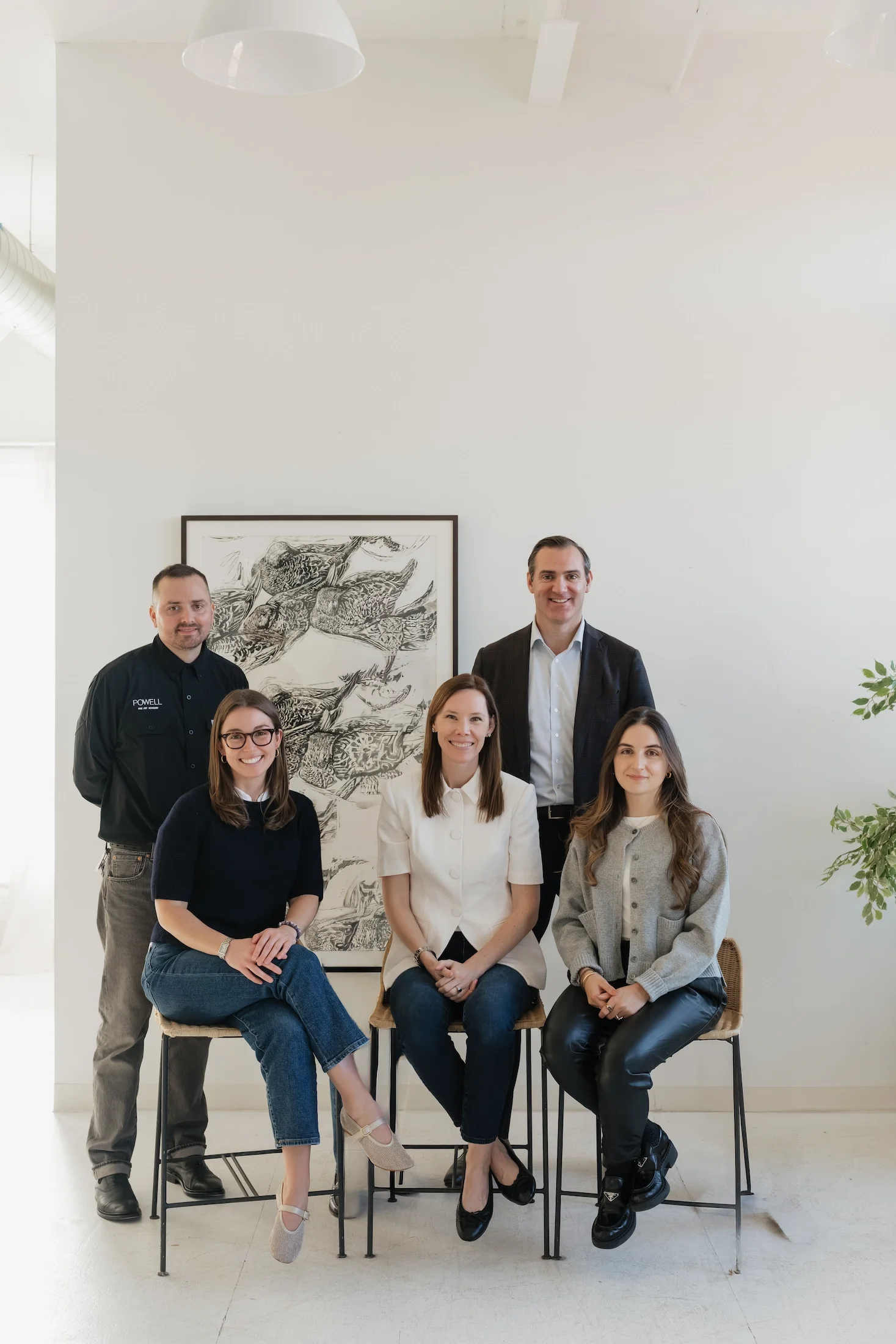A group of seven people, four sitting and three standing, inside a modern room with white walls and minimalist art, smiling at the camera.