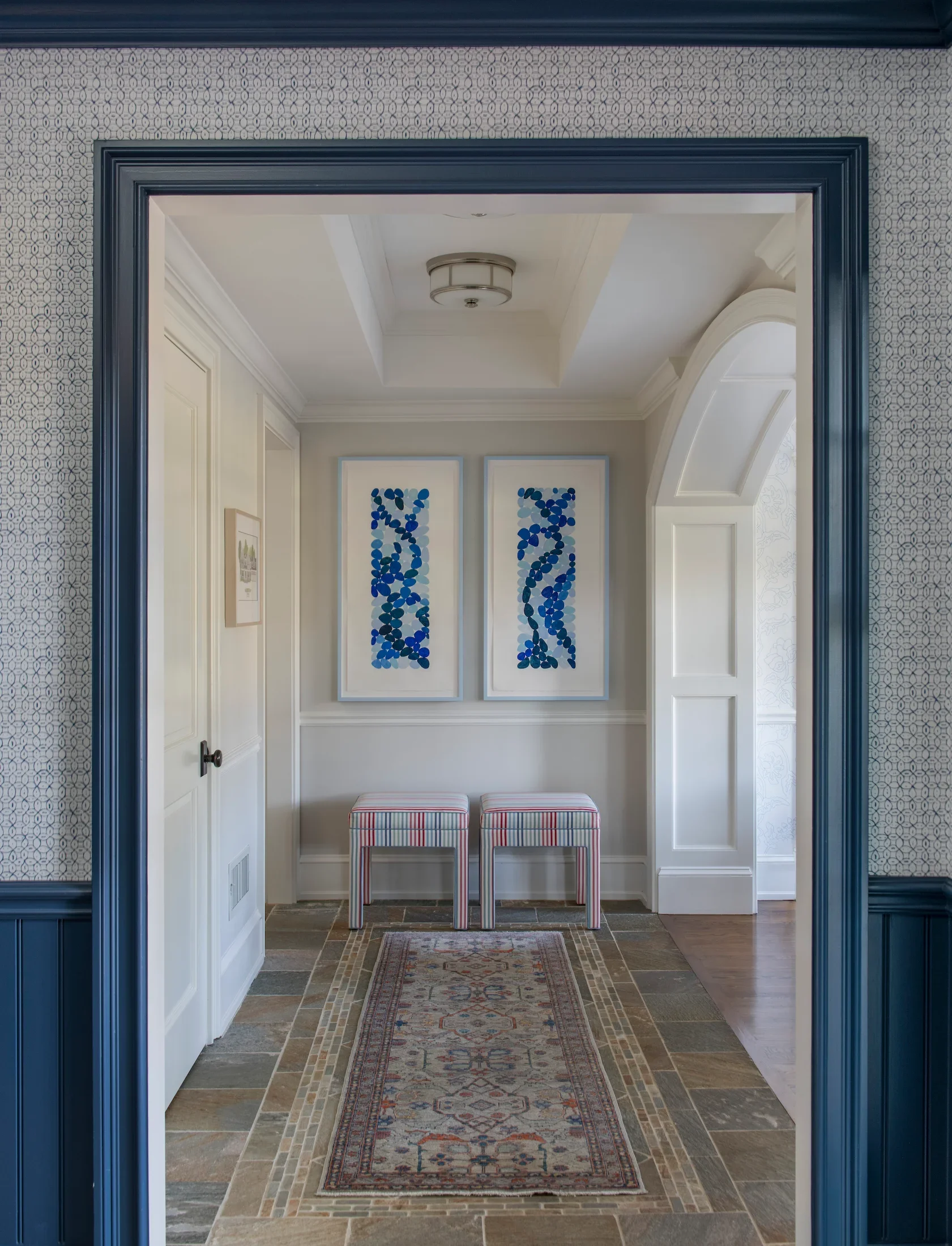 A view through a doorway into a hallway with white walls, featuring two framed abstract blue and black art pieces, two striped stools, and a decorative rug on a tiled floor.