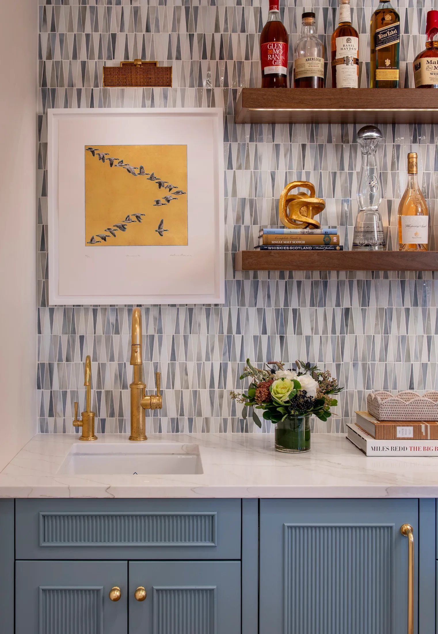 A kitchen countertop with a gold faucet, a small sink, a vase of flowers, and several books, with blue cabinets below. Above, two wooden shelves hold drinks, glassware, and decorative items. The wall has a geometric tile pattern, and a framed art piece of flying birds against a yellow background is on the wall.