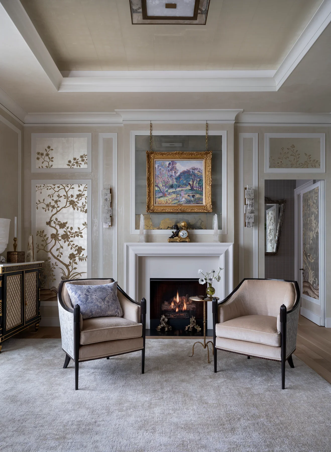 Living room with a fireplace, two beige chairs with black frames and light-colored cushions, a small round table with a flower vase, a framed landscape painting above the fireplace, decorative wall panels with floral patterns, and a carpeted floor.