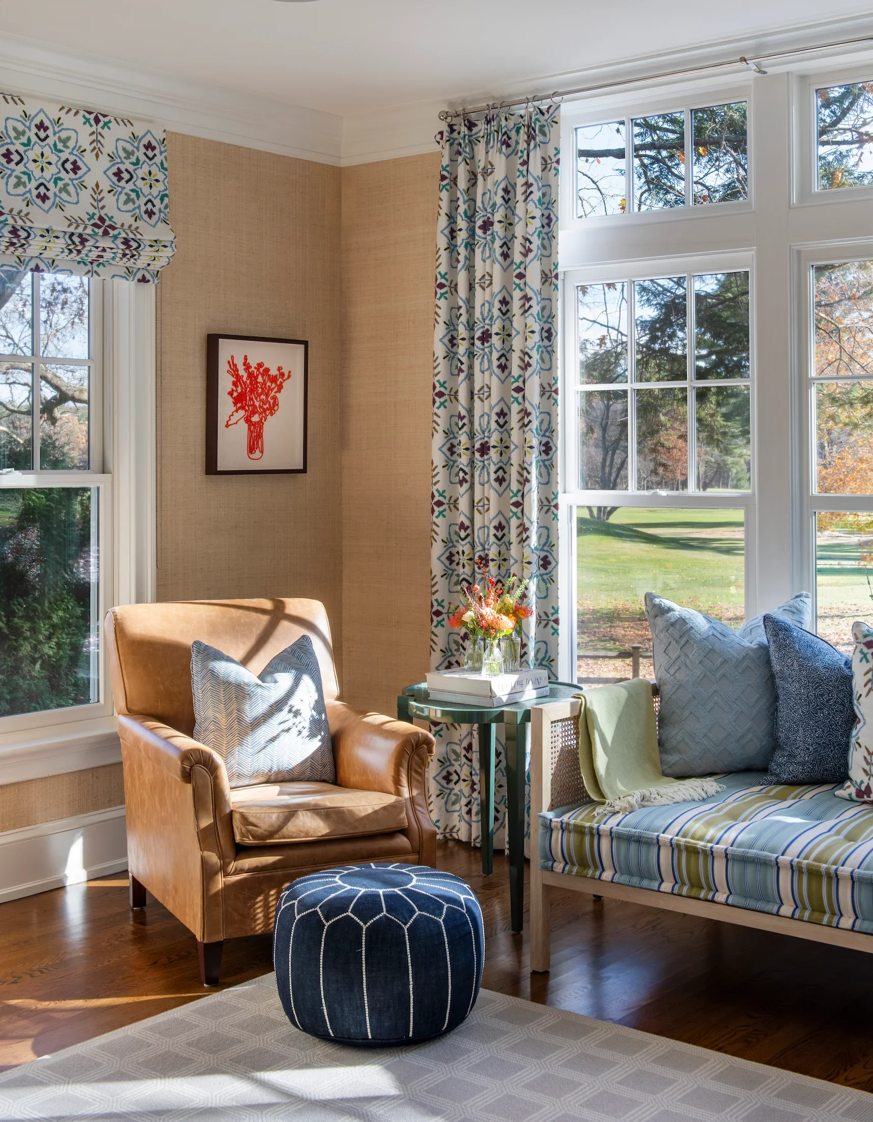 Living room with large windows showing trees outside, brown leather armchair with patterned pillow, blue and white patterned ottoman, striped daybed with pillows and a yellow blanket, side table with flowers, framed art on beige wall, curtains with blue, purple, and brown design.
