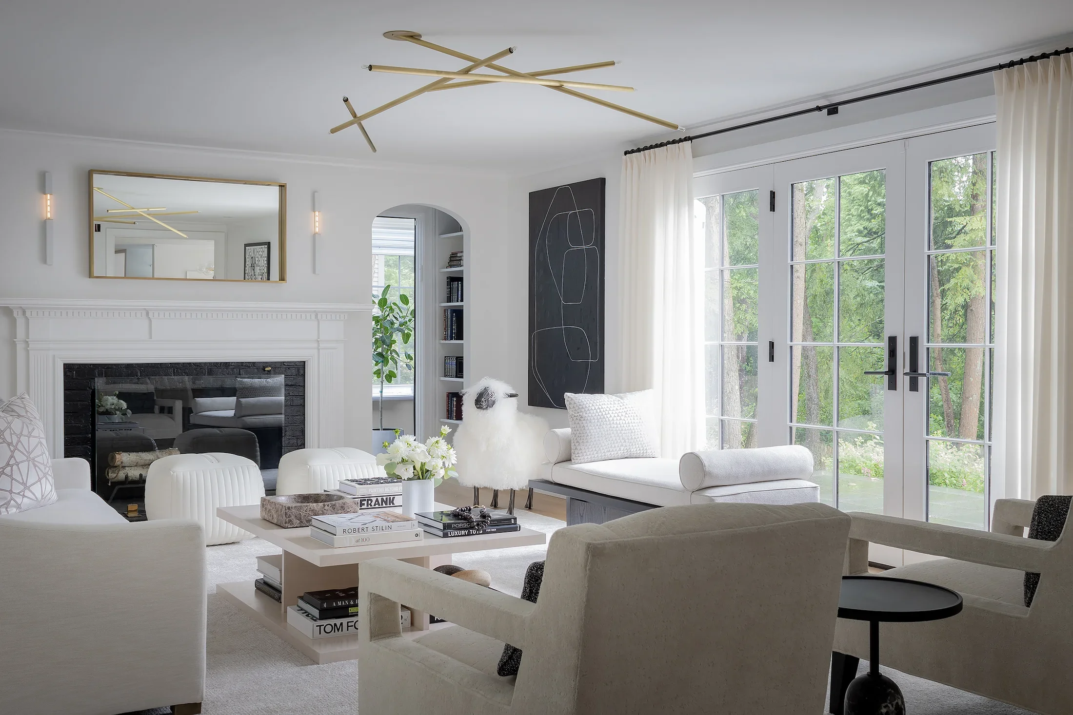 A modern bright living room with white walls, large windows, and white curtains. There are white and light gray furniture pieces, including sofas and chairs, and a coffee table with books and decorative objects. A black fireplace with a mantel is visible, along with a wall mirror and a bookshelf. A ceiling light fixture with intersecting beige rods extends across the ceiling.