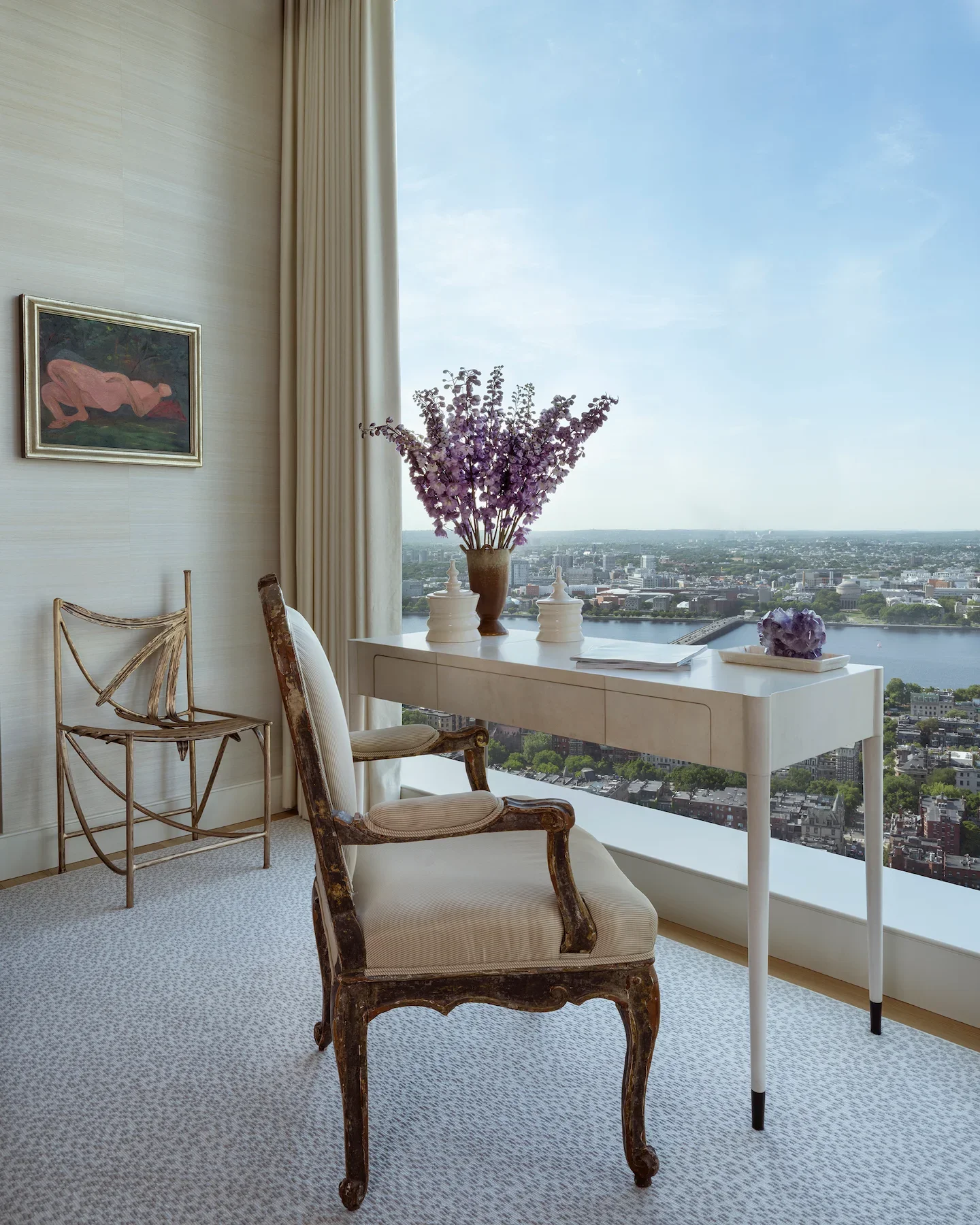 The image shows a corner of a room with a view of a cityscape and river through a large window. The room contains a white desk with decorative items, including a large vase of purple flowers, two white vases, and a purple rock. There are two upholstered chairs with a vintage style, one with worn wood and the other with patchy fabric. The walls are decorated with a framed painting of a pink figure and have beige curtains.