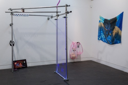 An art gallery. A structure made of pipes dominates the space. Two laundry baskets, pink, hang on the wall, as does a piece of cloth with a work of Larry Stylinson photomanipulation fan art.