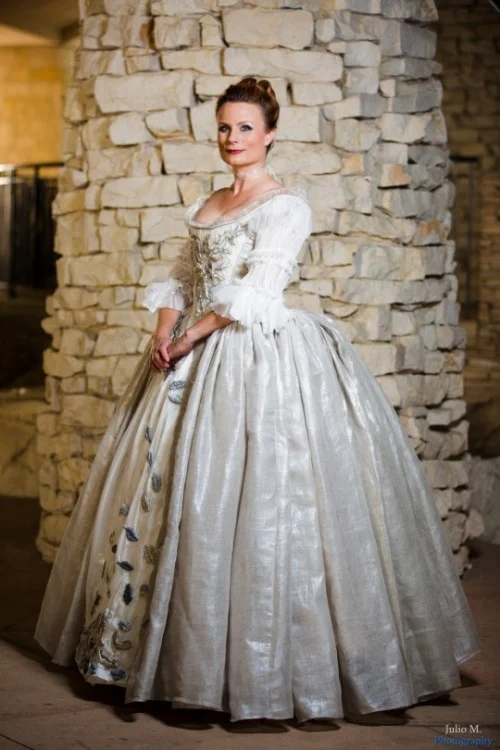 Teresa wearing the wedding dress from Outlander —a pearl silk gown with embroidered metallic leaves all down the bodice and a very large and sweeping skirt—posing in front of a stone wall.