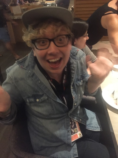 Britta, wearing glasses, a snapback and button-down denim shirt, grins and waves at the camera.
