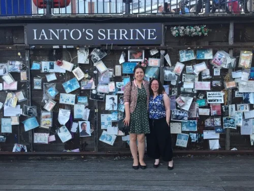 Elizabeth and Dr. Naomi Jacobs standing in front of Ianto’s Shrine, a wall covered in flyers and photographs fans have tacked up commemorating Ianto from Torchwood .