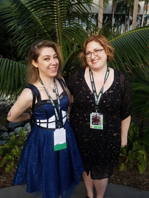 Katytastic and Alexa Donne pose in front of greenery. Katy wears a TARDIS dress and Alexa a spangled black dress.