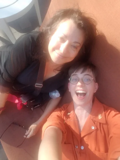 Annie Nocenti and Flourish pose on a sofa, holding the camera above their heads. It is very sunny.