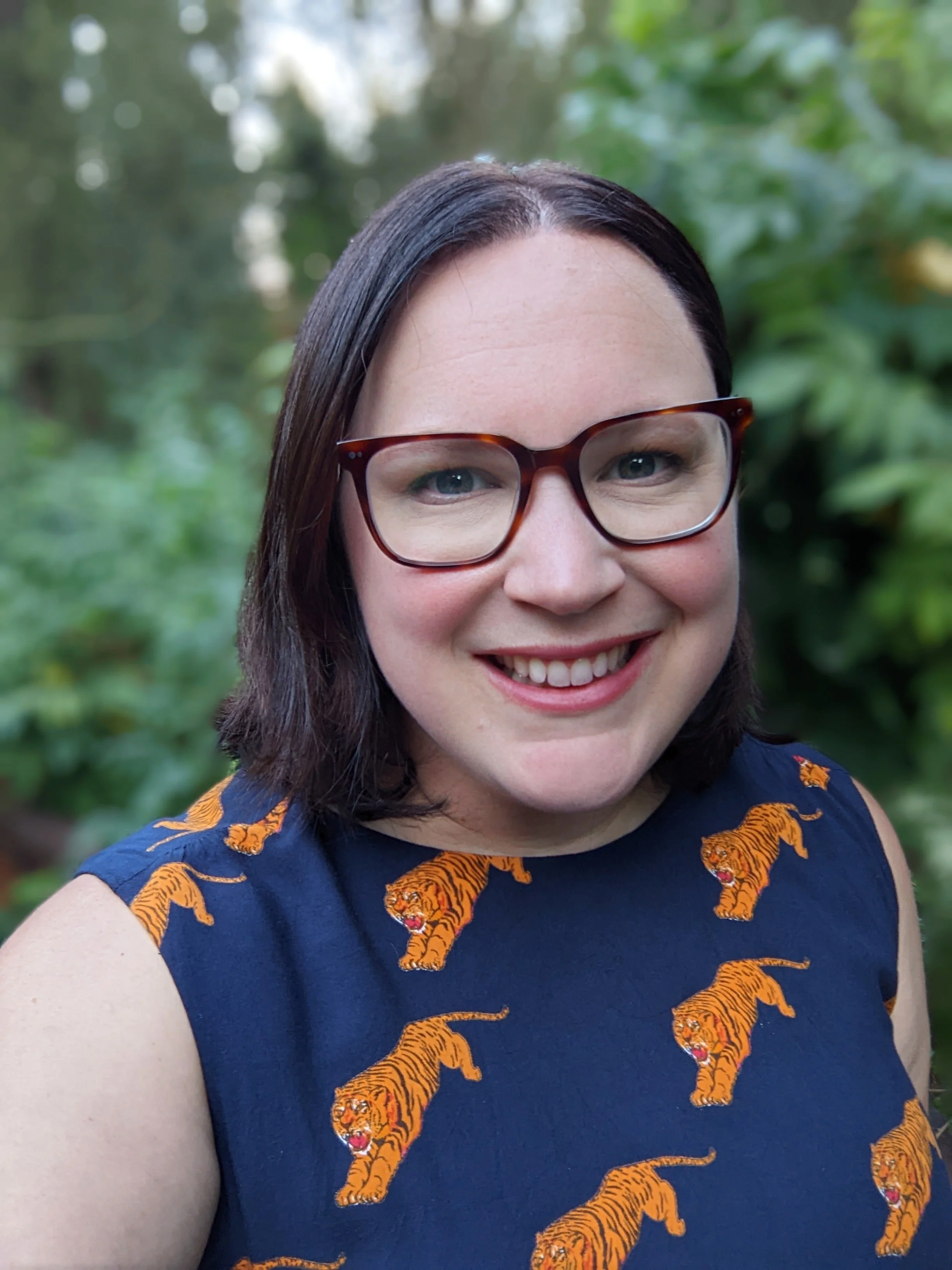Headshot of Laura: fair-skinned and dark-haired, wearing glasses and a navy dress with orange tigers printed on it.