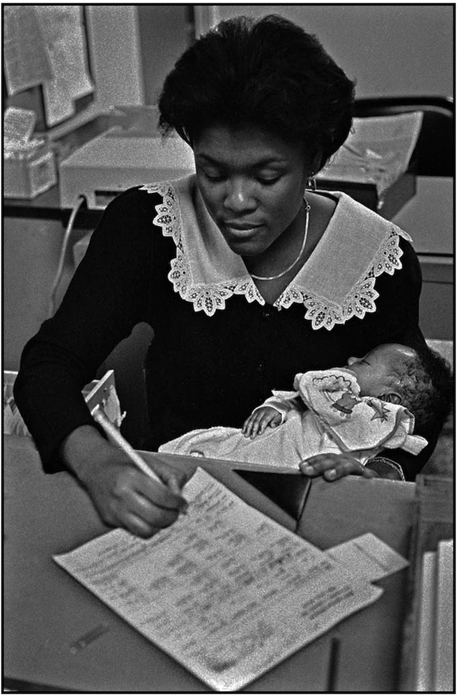   Office Aide Kettly Aime does her work while caring for child during mother's &nbsp;examination at Kings County Hospital. 1988.  