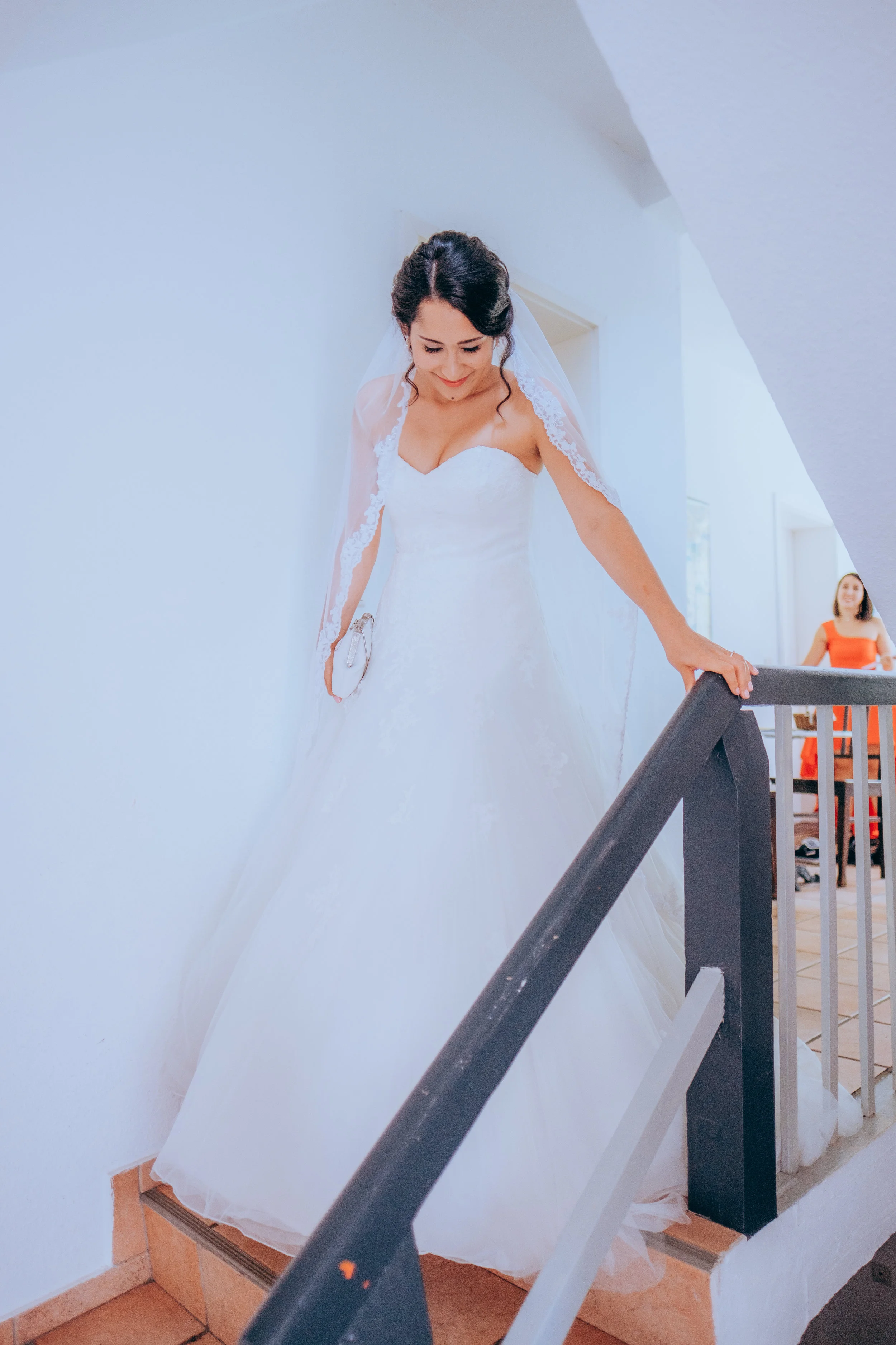 Braut in einem weißen Hochzeitkleid, die auf einer Treppe steht, mit einem Schleier auf dem Kopf, im Hintergrund eine Frau in einem roten Kleid.