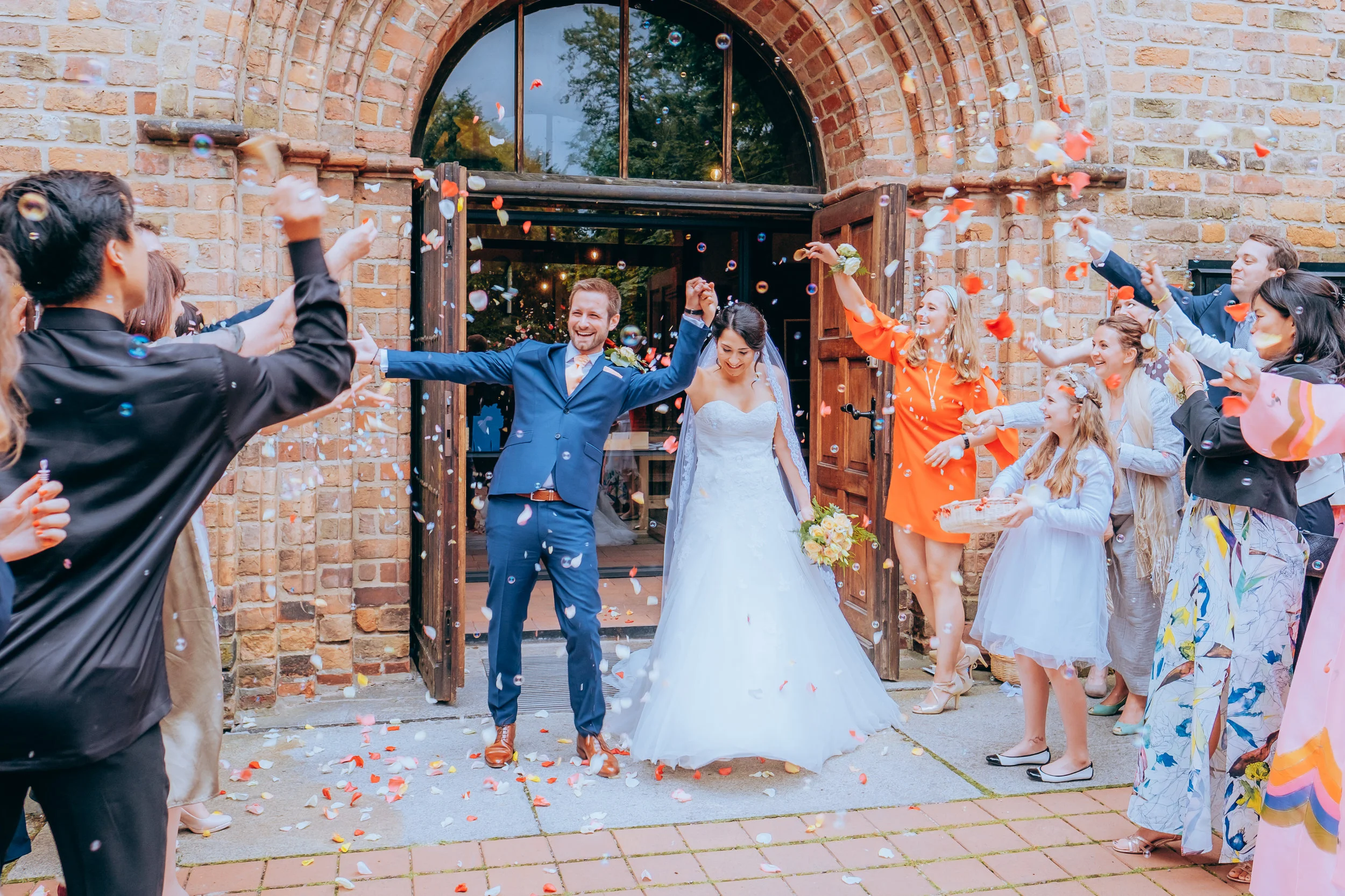 Hochzeitspaar, das aus einer alten Steintür tritt, umgeben von Freunden, die Konfetti werfen, vor einer Backsteinwand.