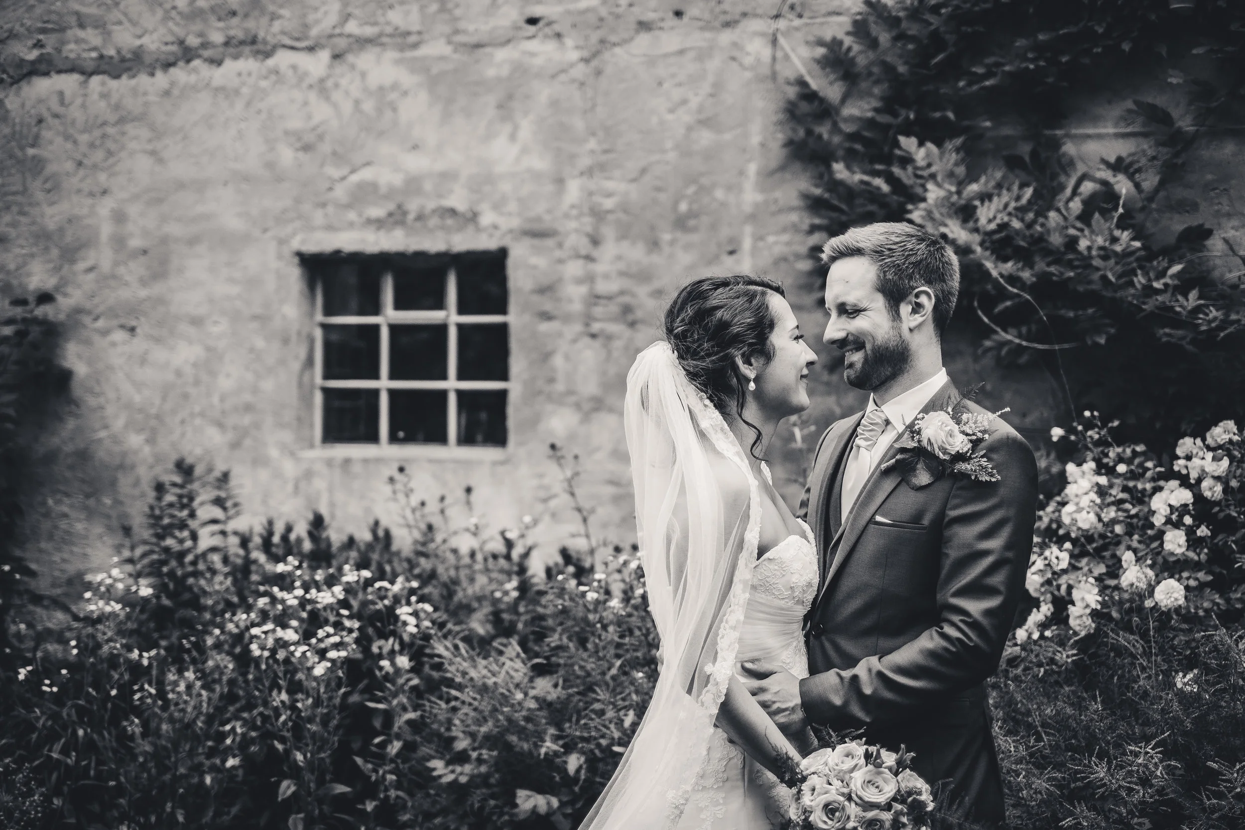 Ein Brautpaar steht sich liebevoll gegenüber vor einer alten Steingebäude, umgeben von Gartenblumen, bei einer Hochzeit im Freien, in Schwarz-Weiß fotografiert.