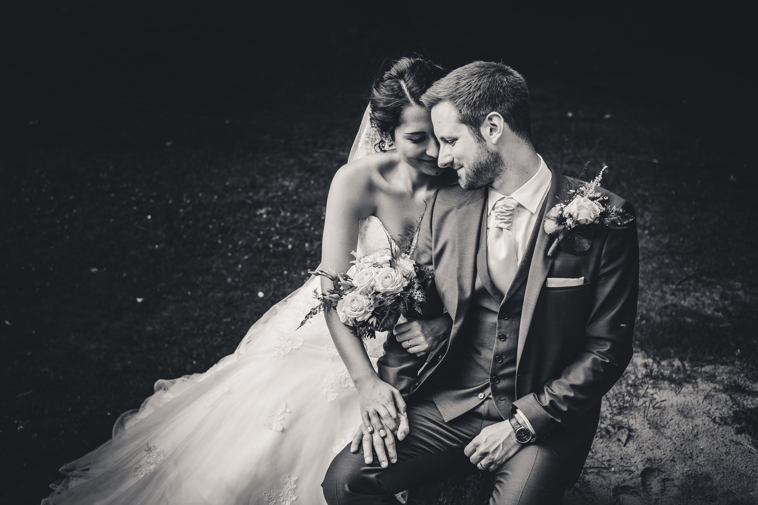 Schwarz-weiß-Foto eines Paares bei der Hochzeit, das sich an die Stirn fasst und lächelt, mit Blumen. Der Hintergrund ist dunkel.