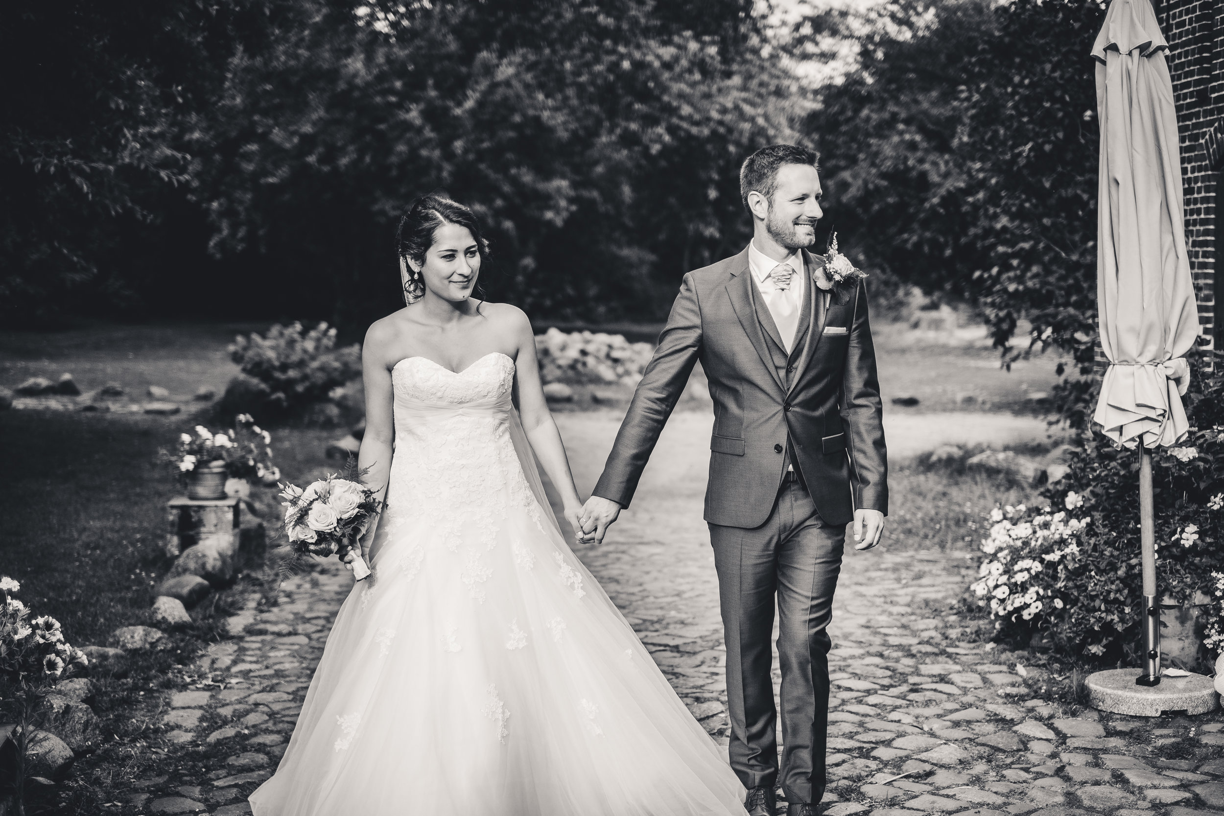 Ein schwarz-weiß-Bild eines verheirateten Paares bei ihrer Hochzeit, das Hand in Hand auf einer Gartenstraße spaziert, umgeben von Blumen und Bäumen.