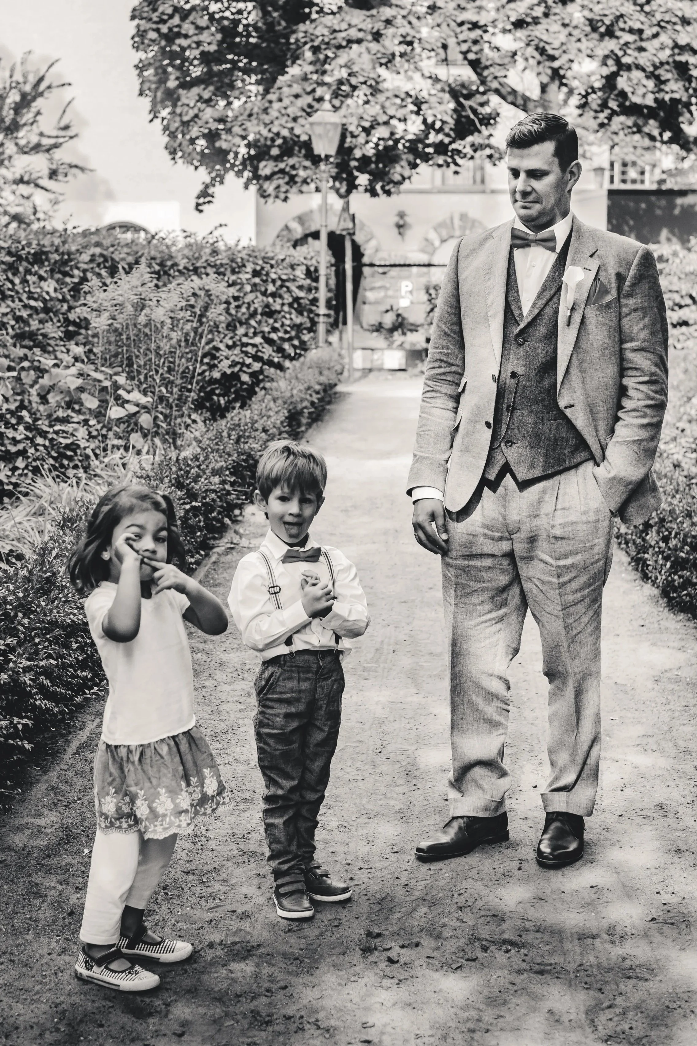 Schwarz-weiß-Foto eines Mannes in formeller Kleidung mit Anzug, Weste und Fliege, der mit drei Kindern auf einem Gartenweg geht. Die Kinder tragen legere Kleidung mit Hemd, Rock und Hose, und lachen oder schauen zur Kamera. Im Hintergrund Bäume und ein Gebäude.