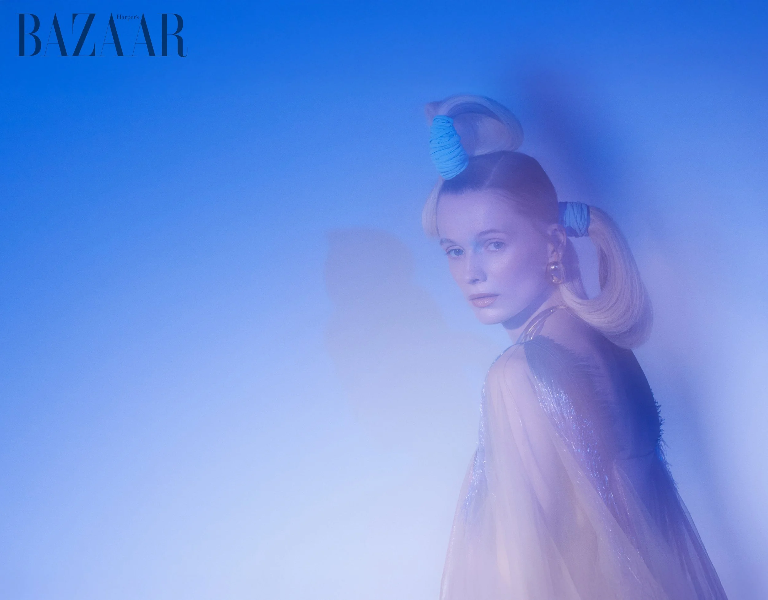 A woman with light skin and blonde hair styled with blue and white striped hair rollers. The photo has a blue tinted overlay, and the woman is looking at the camera with a neutral expression. The image is branded with the Harvard's Harper's Bazaar logo in the top left corner.
