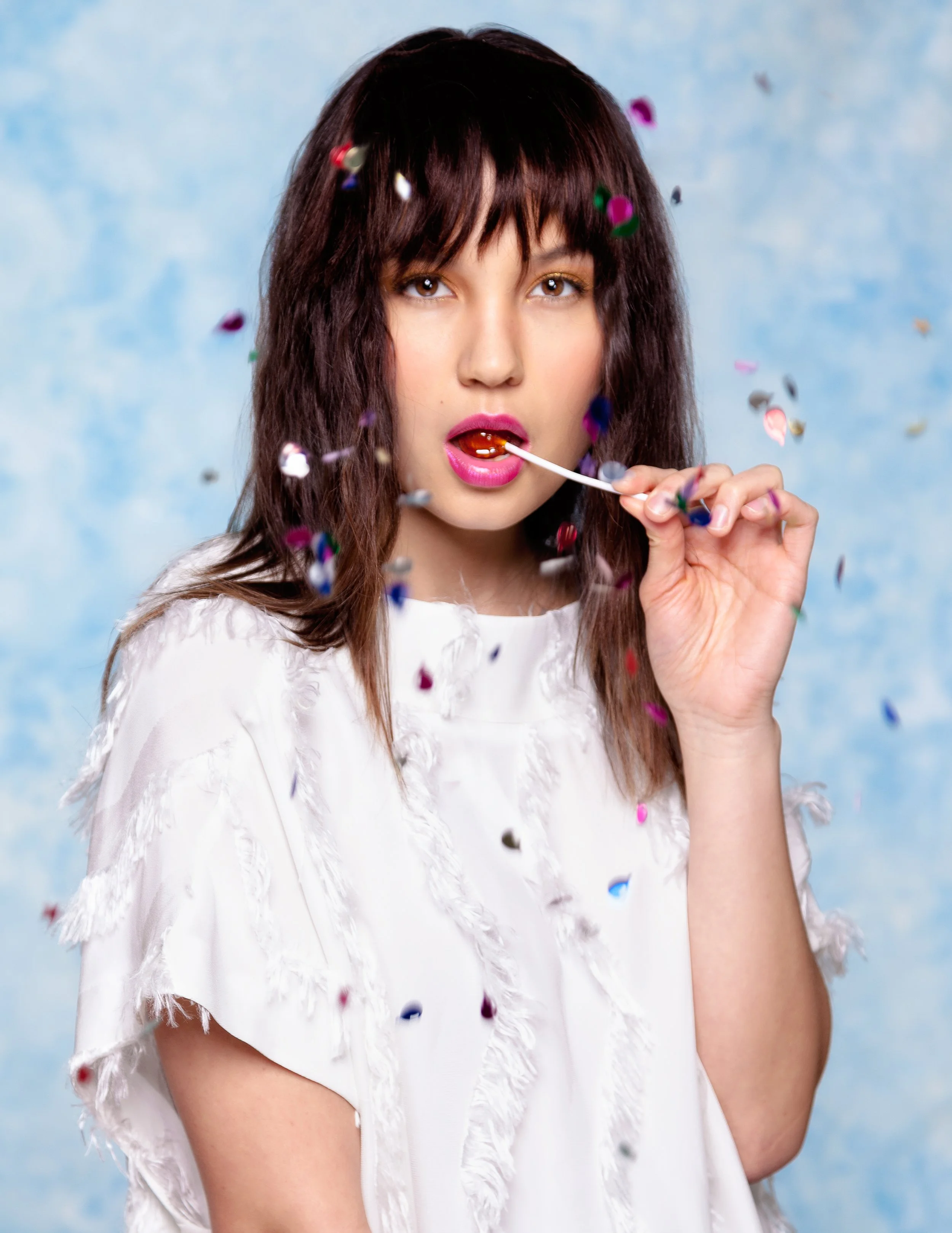 A woman with brown wavy hair and light makeup, wearing a white feathered top, is surrounded by falling colorful confetti. She is holding a lollipop near her lips against a light blue background.