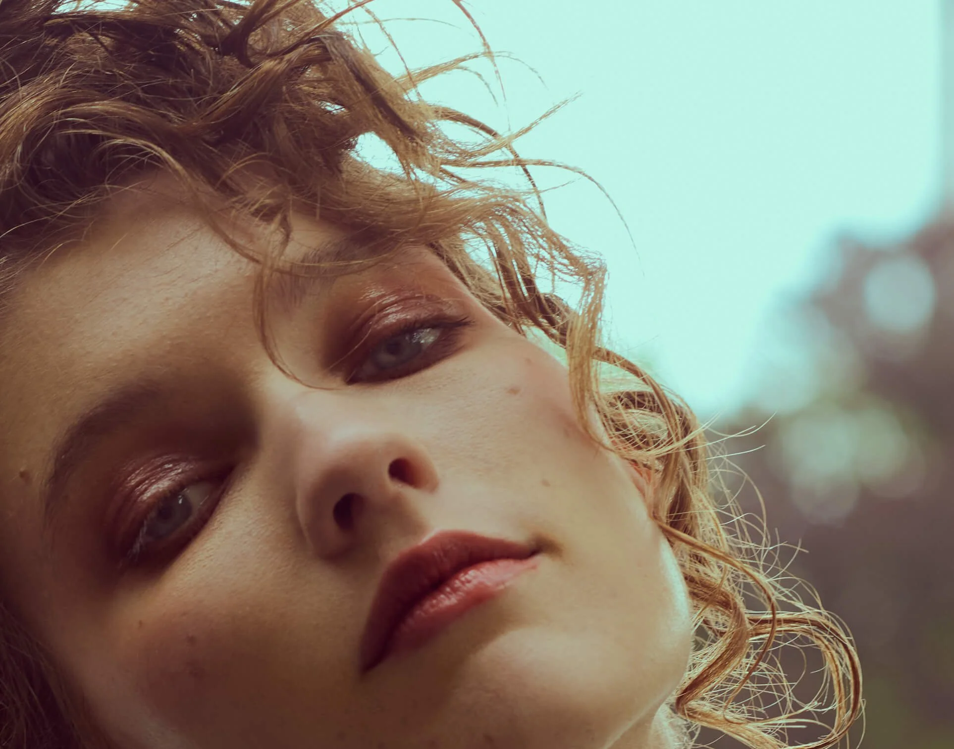 Close-up of a woman's face with curly red hair, light skin, gray eyes, full lips, and subtle makeup, with a blurred outdoor background.