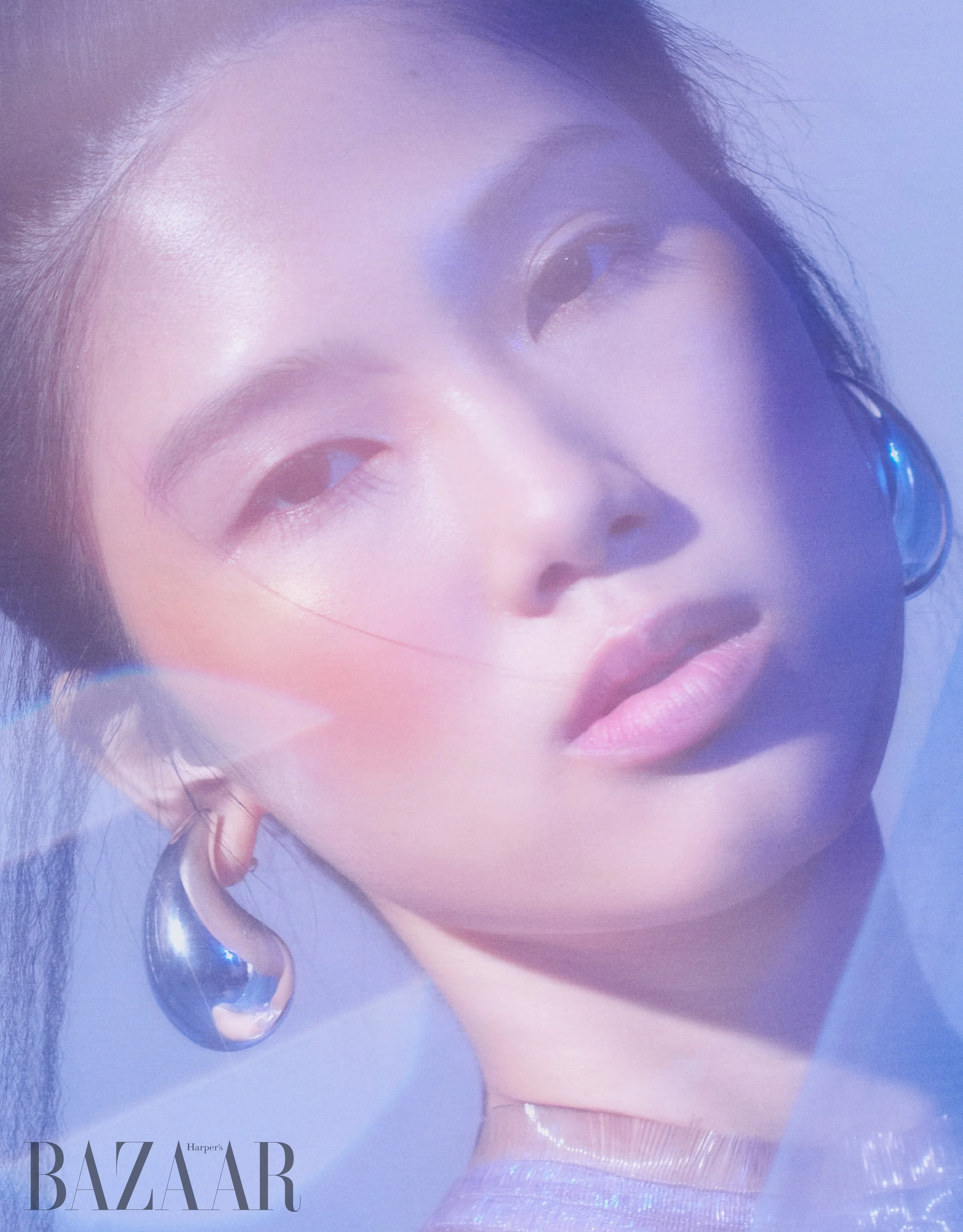 Close-up of a woman's face with makeup, with her eye partially visible, wearing earrings, and a lavender-colored garment. The photo appears to be from a magazine cover of Harper's Bazaar.