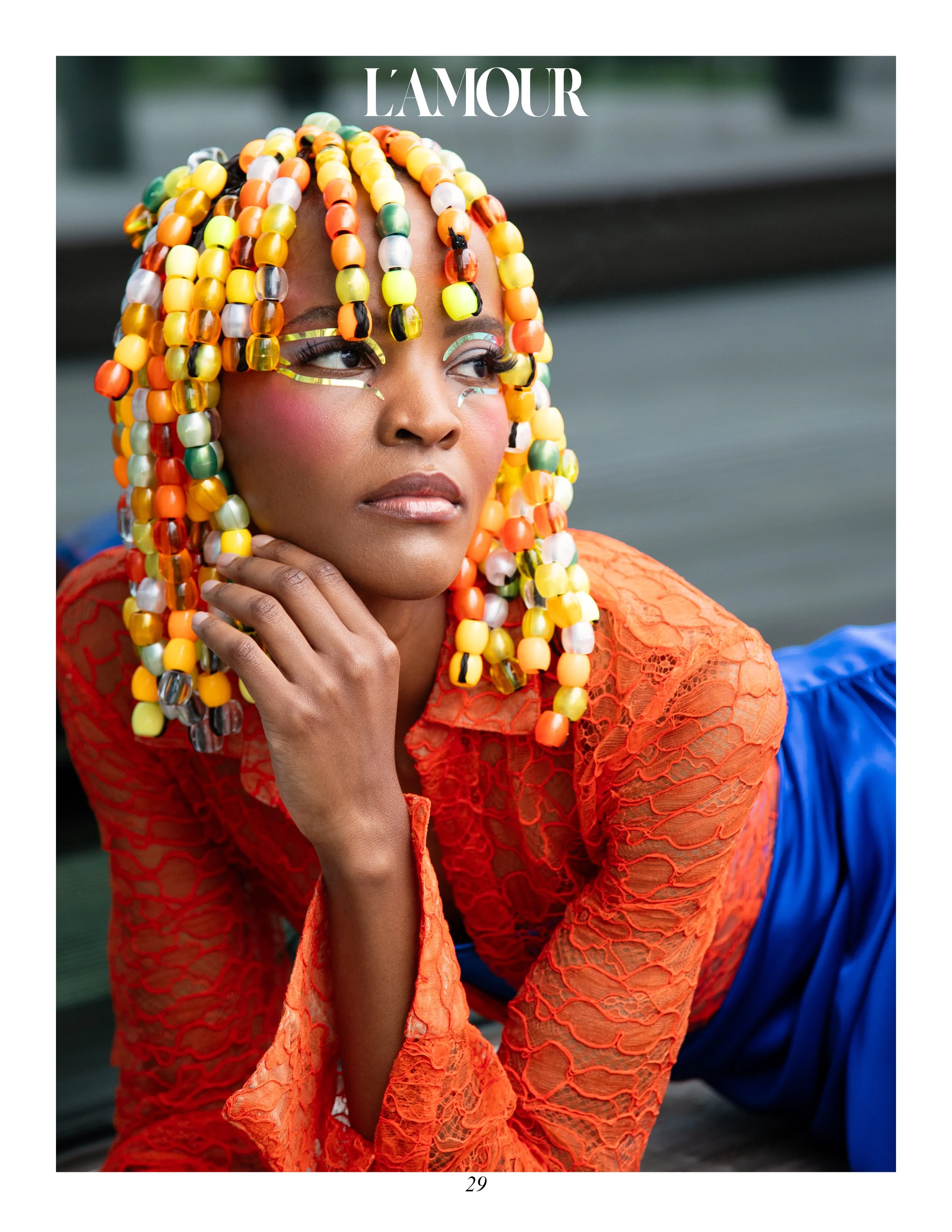 A woman with colorful beaded hair, wearing eye-catching makeup, an orange lace top, and blue pants, seated outdoors with a blurred background.
