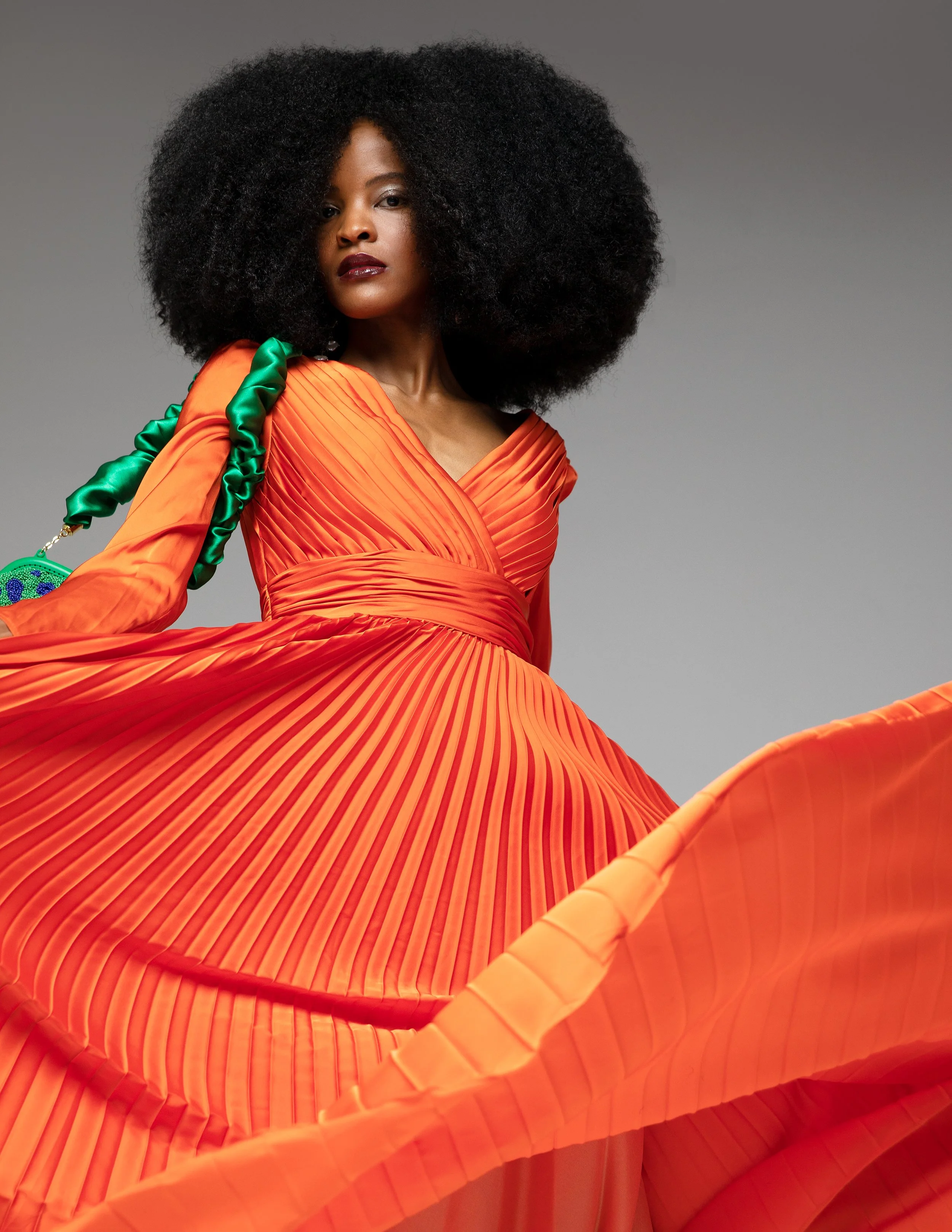 A woman with dark skin and voluminous curly hair, wearing a vibrant orange dress with pleated design and a green satin strap, poses against a plain gray background.