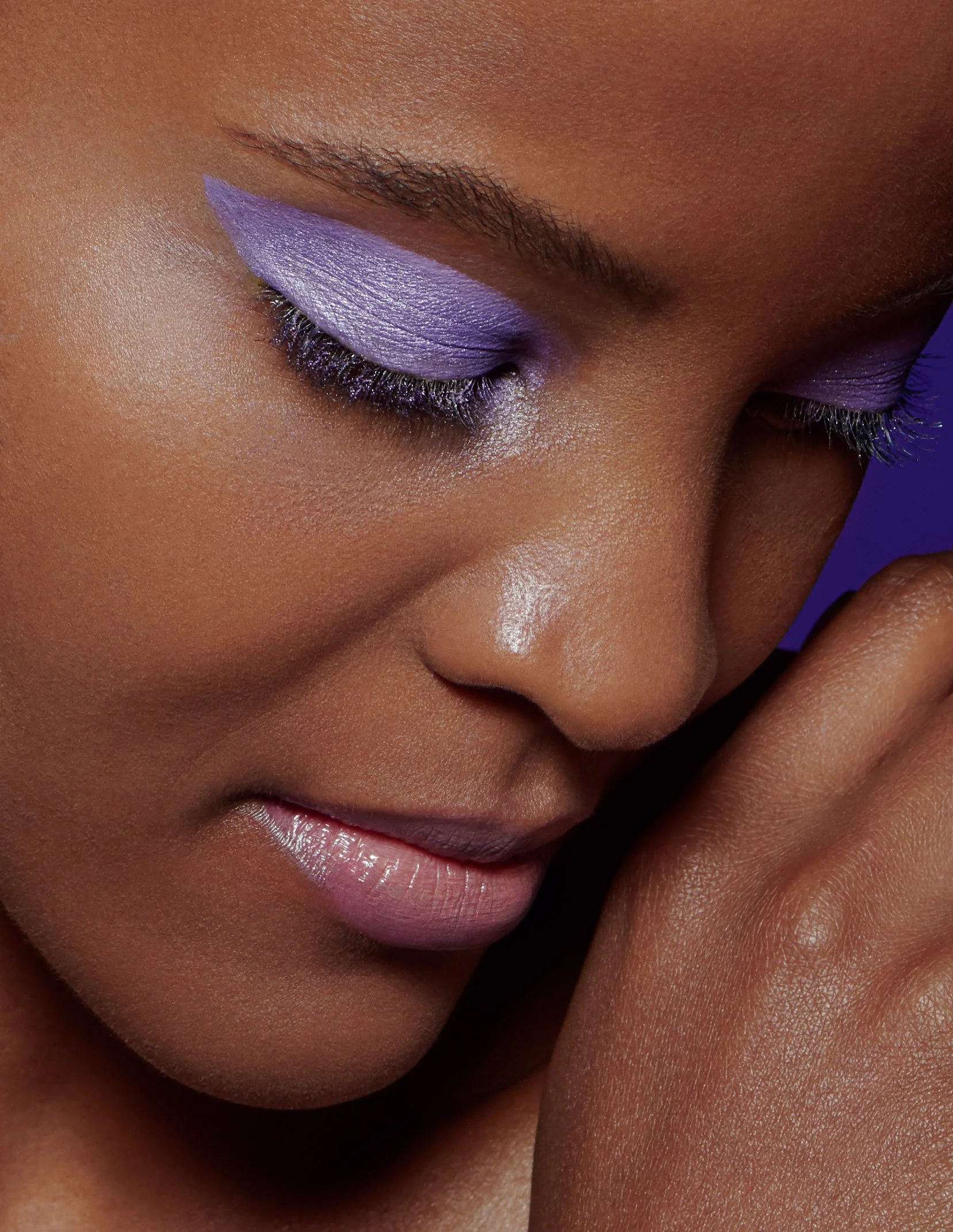 Close-up of a woman's face with purple eyeshadow and glossy lips, her eyes closed and hand near her cheek.