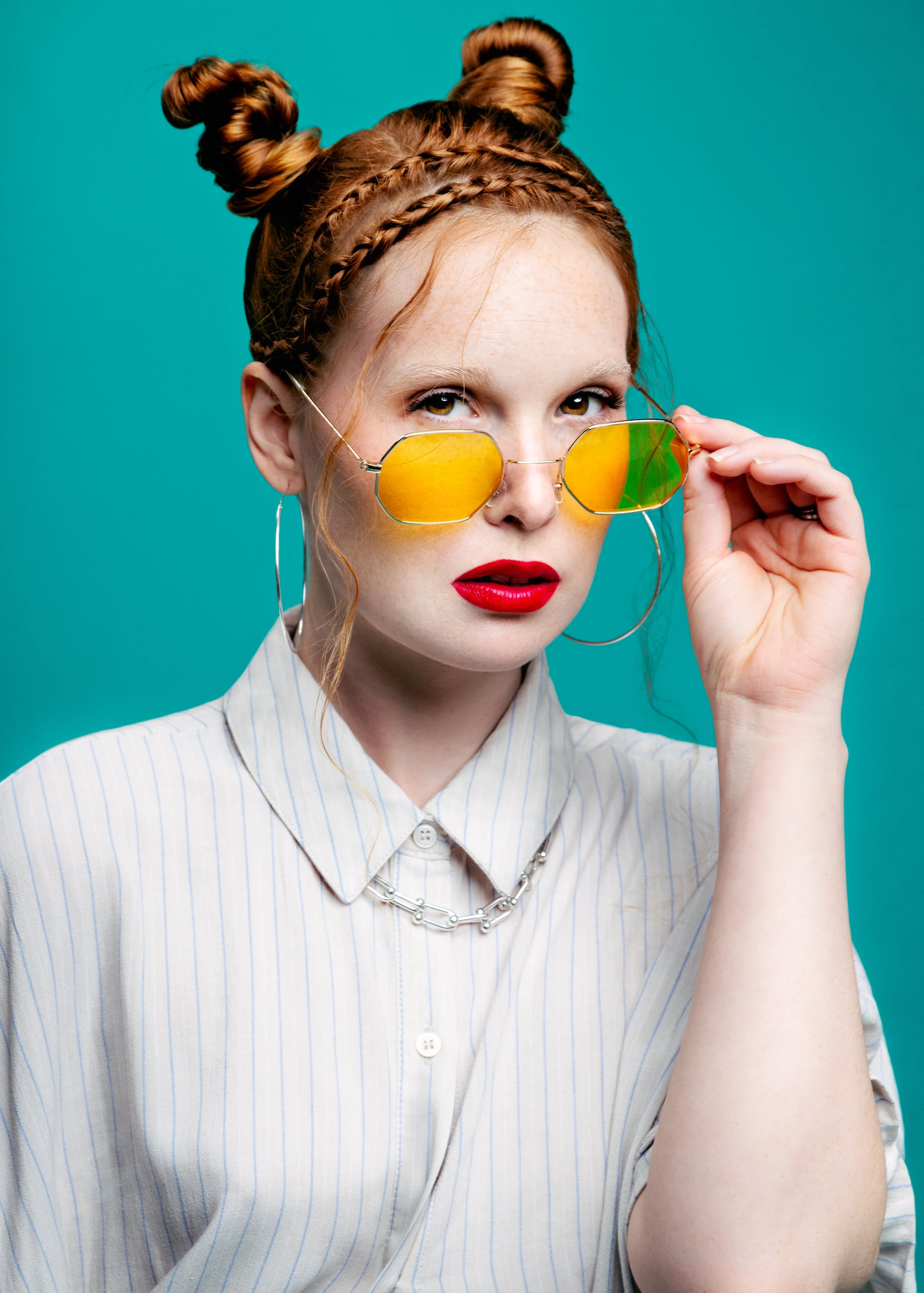 A young woman with red hair styled in space buns and braided details, wearing yellow mirrored sunglasses, red lipstick, hoop earrings, a silver chain necklace, and a light-colored striped button-up shirt, against a teal background.