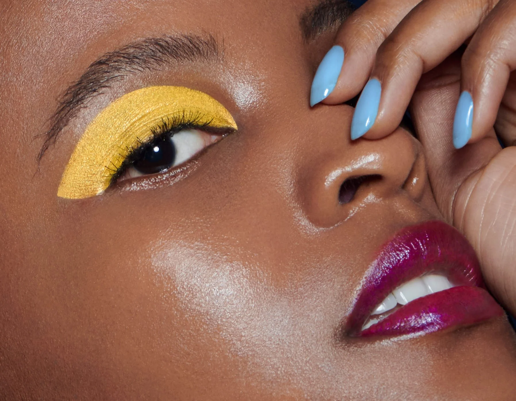 Close-up of a woman's face with makeup, including bright yellow eyeshadow, glossy pink lipstick, and a partially visible smile. Her eye is decorated with glitter and she has manicured nails painted in a light blue color.
