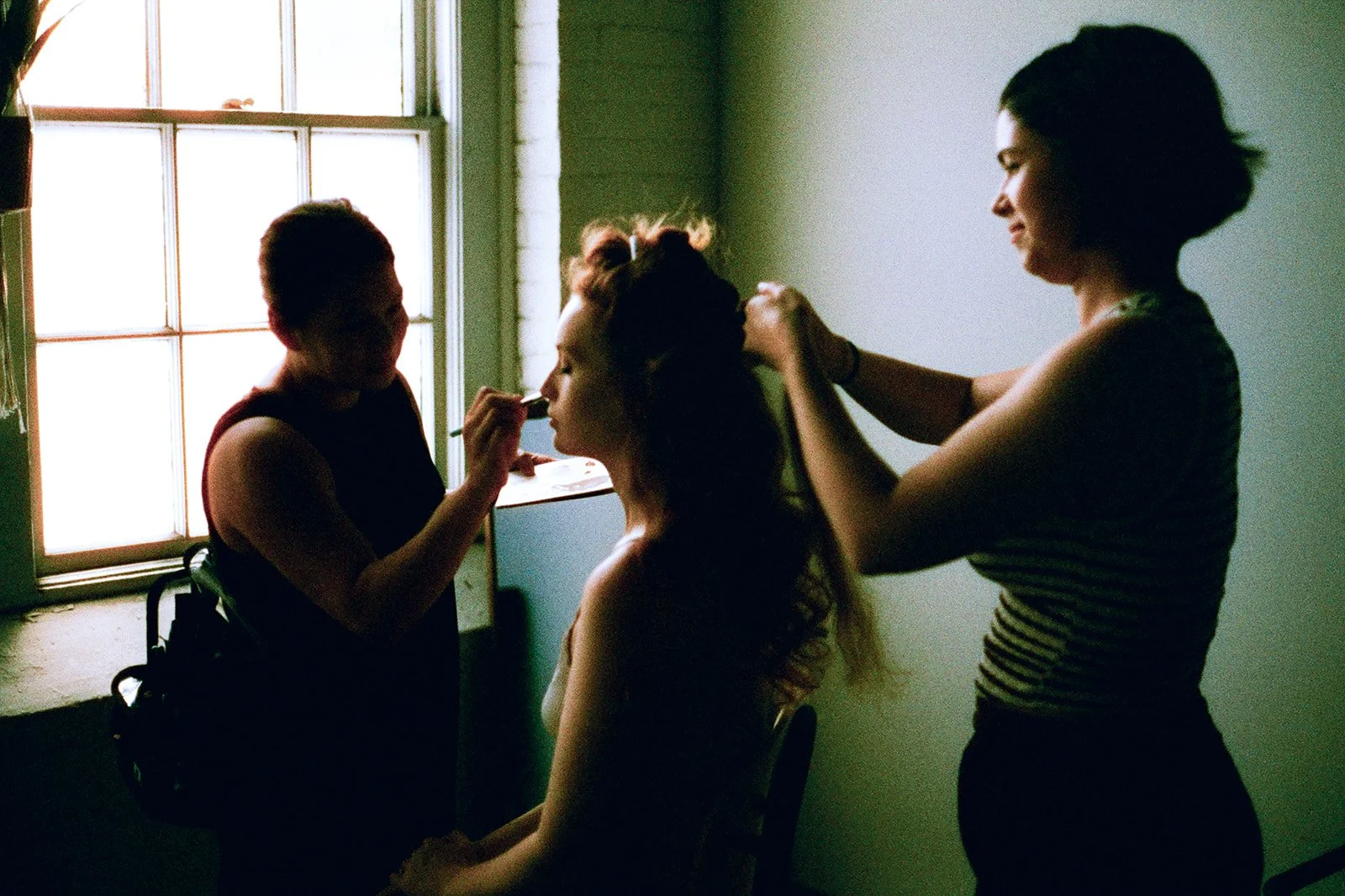 A woman receiving a makeup application from a makeup artist, with another woman styling her hair, in a softly lit room near a window.