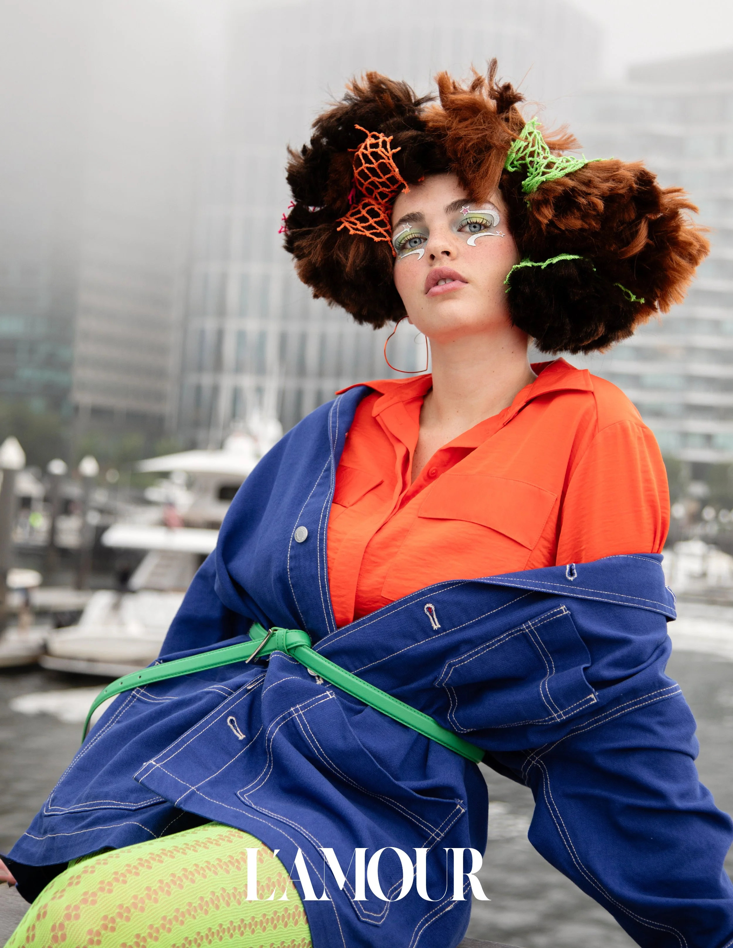 A young woman with curly, voluminous hair adorned with colorful hair net accessories, standing outdoors near a marina with boats, wearing a bright orange shirt, a blue jacket, and yellow patterned leggings, with the words 'L'AMOUR' overlayed in text.