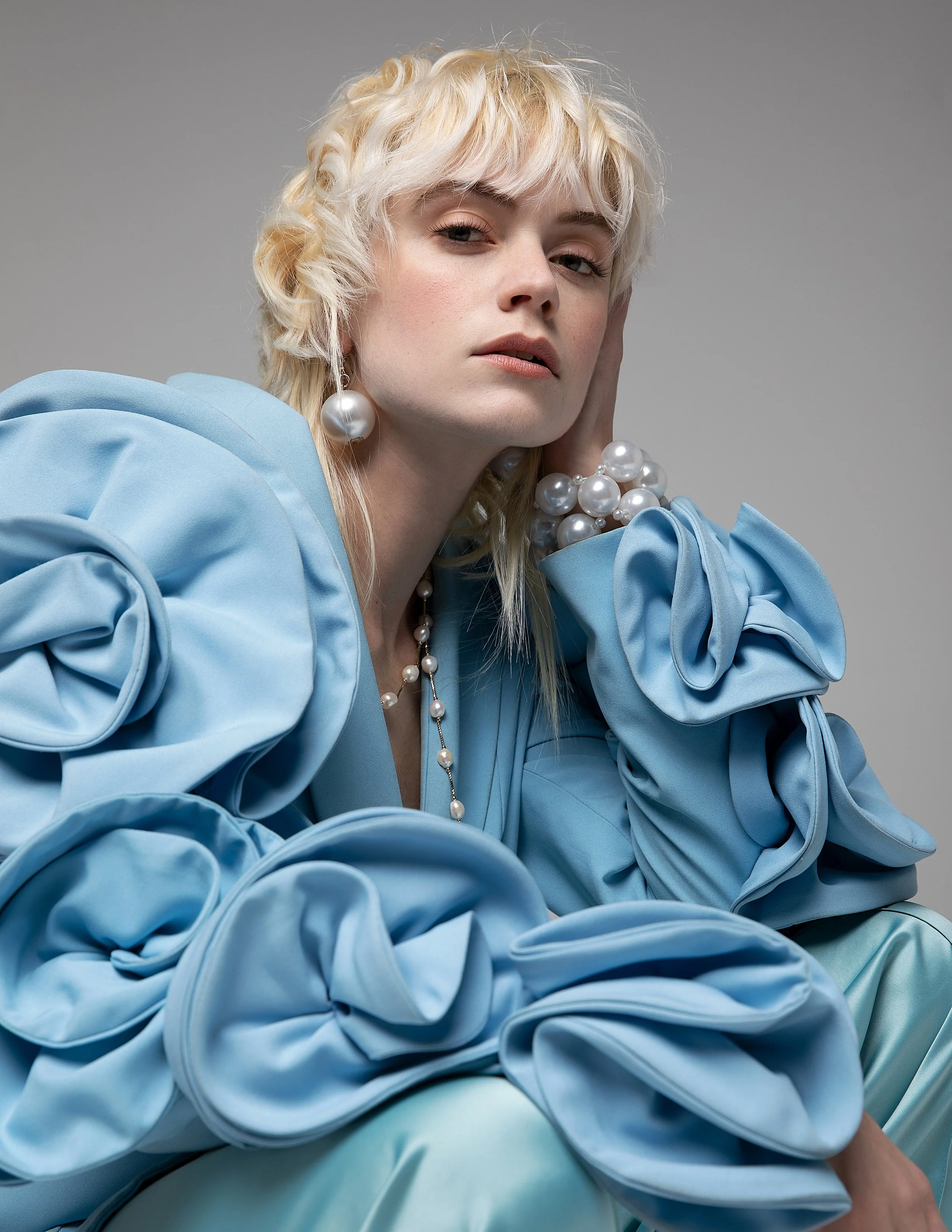 Close-up portrait of a woman with platinum blonde, curly hair, wearing pearl earrings and a pearl necklace, dressed in a blue satin blouse with large ruffled sleeves, against a plain gray background.