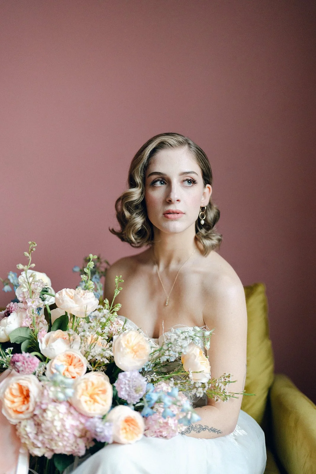 A young woman with short wavy brown hair, wearing a strapless white dress and jewelry, holds a large bouquet of pastel-colored flowers, sitting on a yellow couch against a pink background.