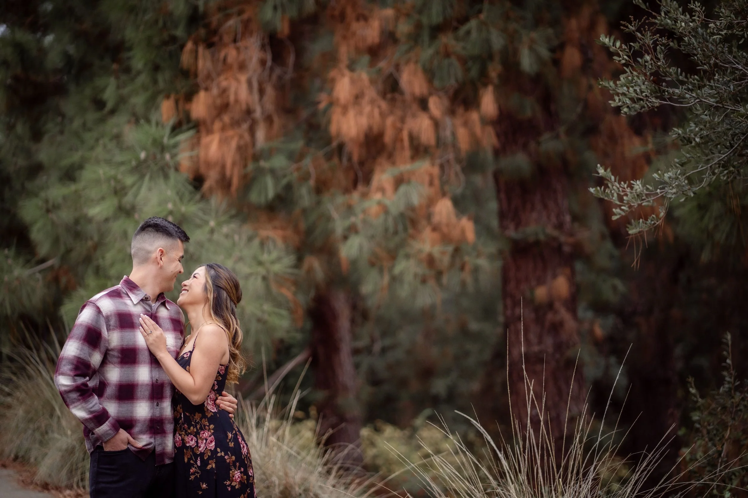 A young couple stands close together outdoors in front of tall trees, smiling and looking into each other's eyes during their engagement session at the UC Davis Arboretum. Photography by Katherine Nicole Photography.