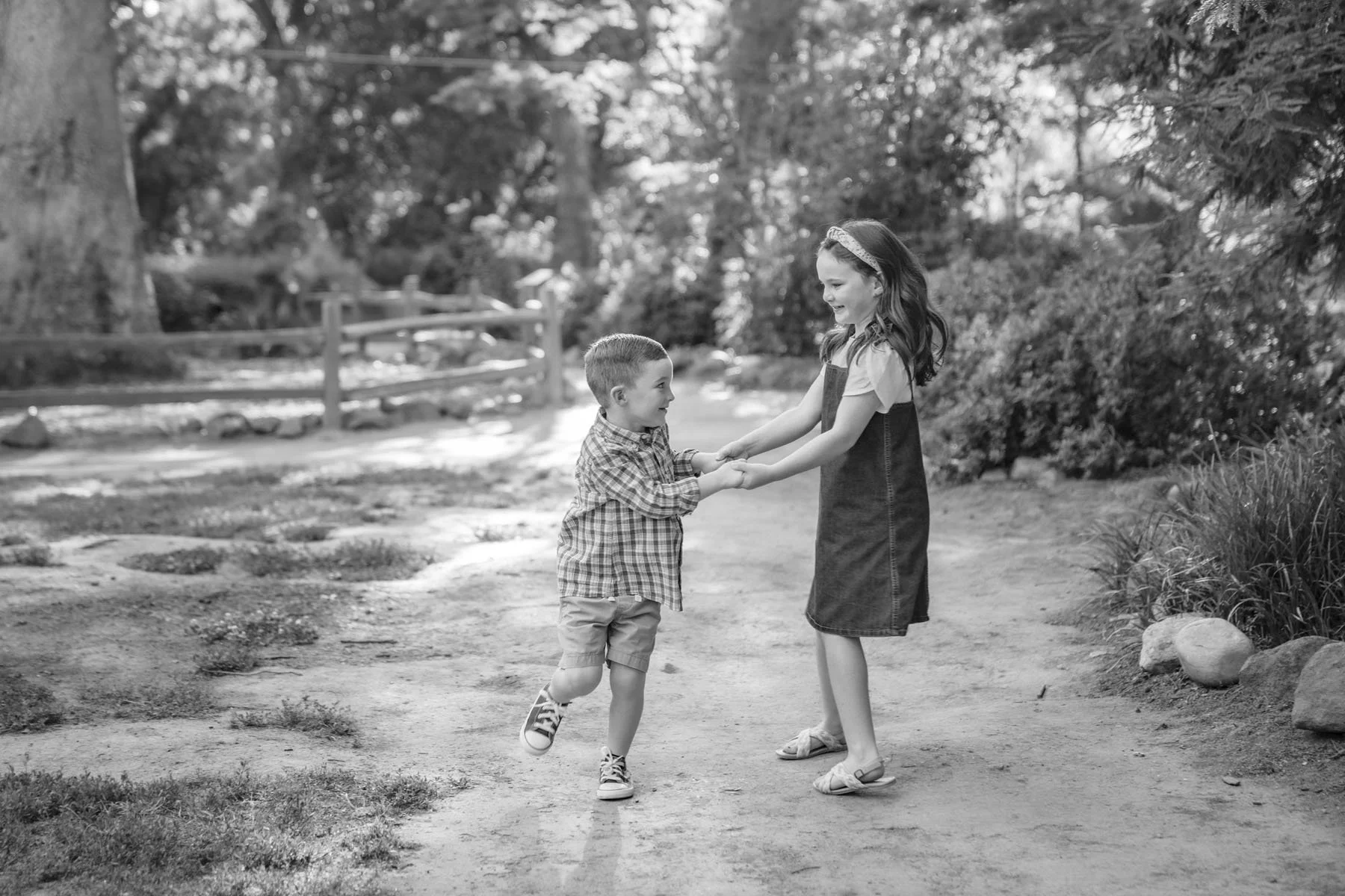 A girl and a boy holding hands and playing outdoors on a dirt path surrounded by trees and bushes, with a wooden fence in the background. Family photos at Jensen Botanical Garden with Sacramento family photographer, Katherine Nicole Photography.