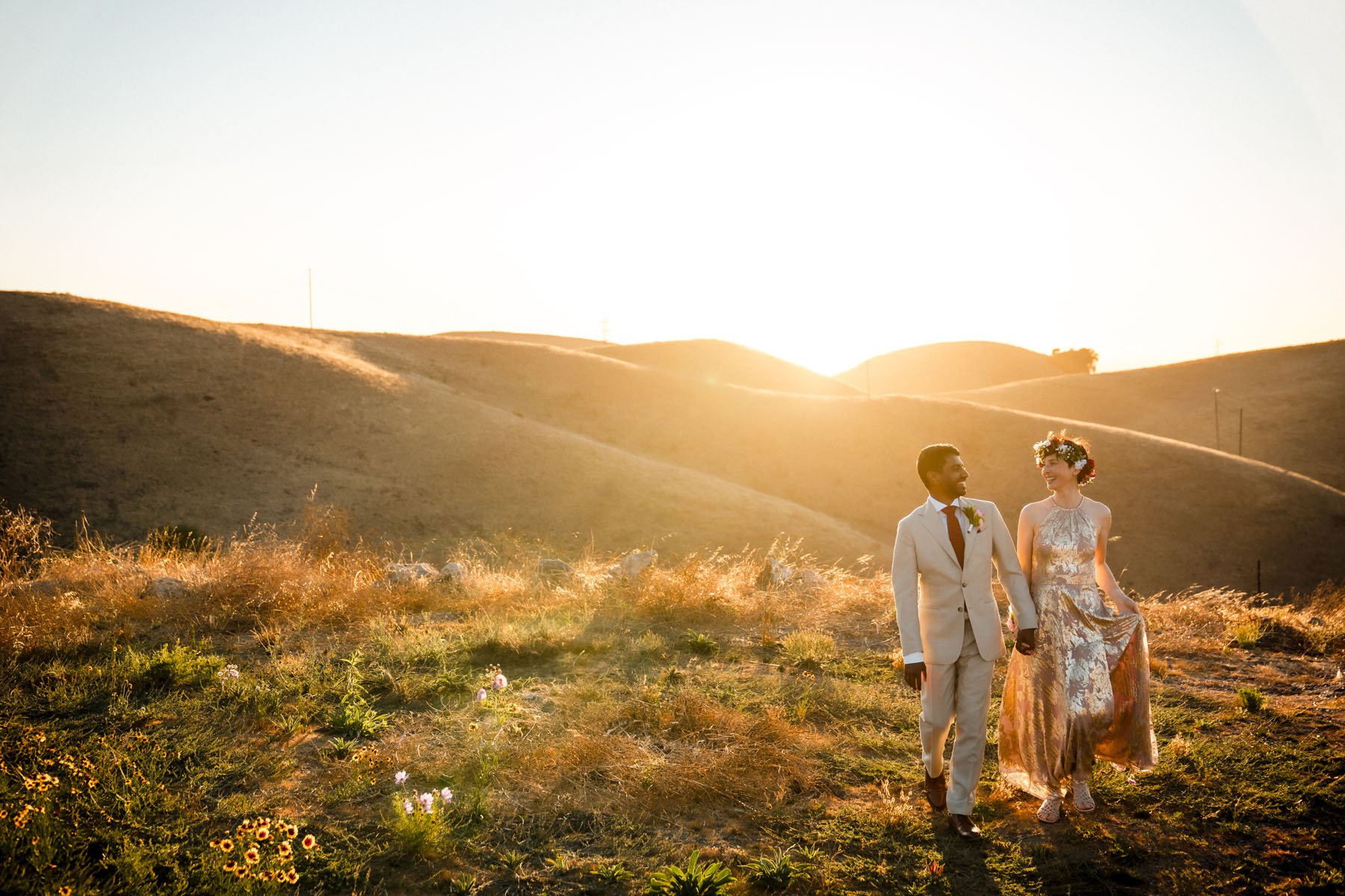 A couple dressed in formal wedding attire walking hand in hand on a grassy hillside at sunset, with rolling hills in the background, during their Nella Terra Cellars wedding. Photography by Sacramento wedding photographer Katherine Nicole Photography