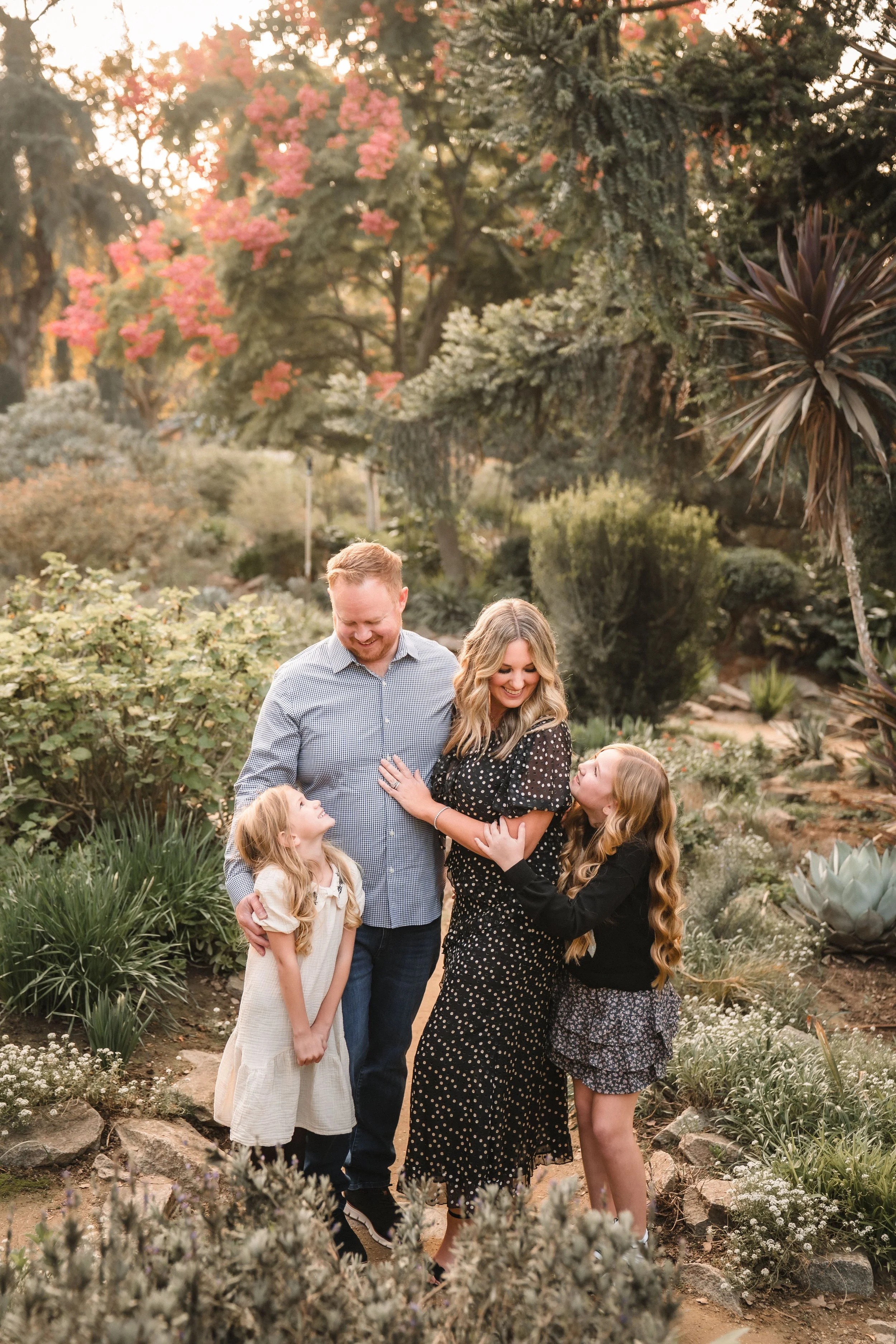A family of five, including two adult women, one adult man, and two young girls, smiling and interacting in a lush garden with various plants, trees, and flowers during sunset.
