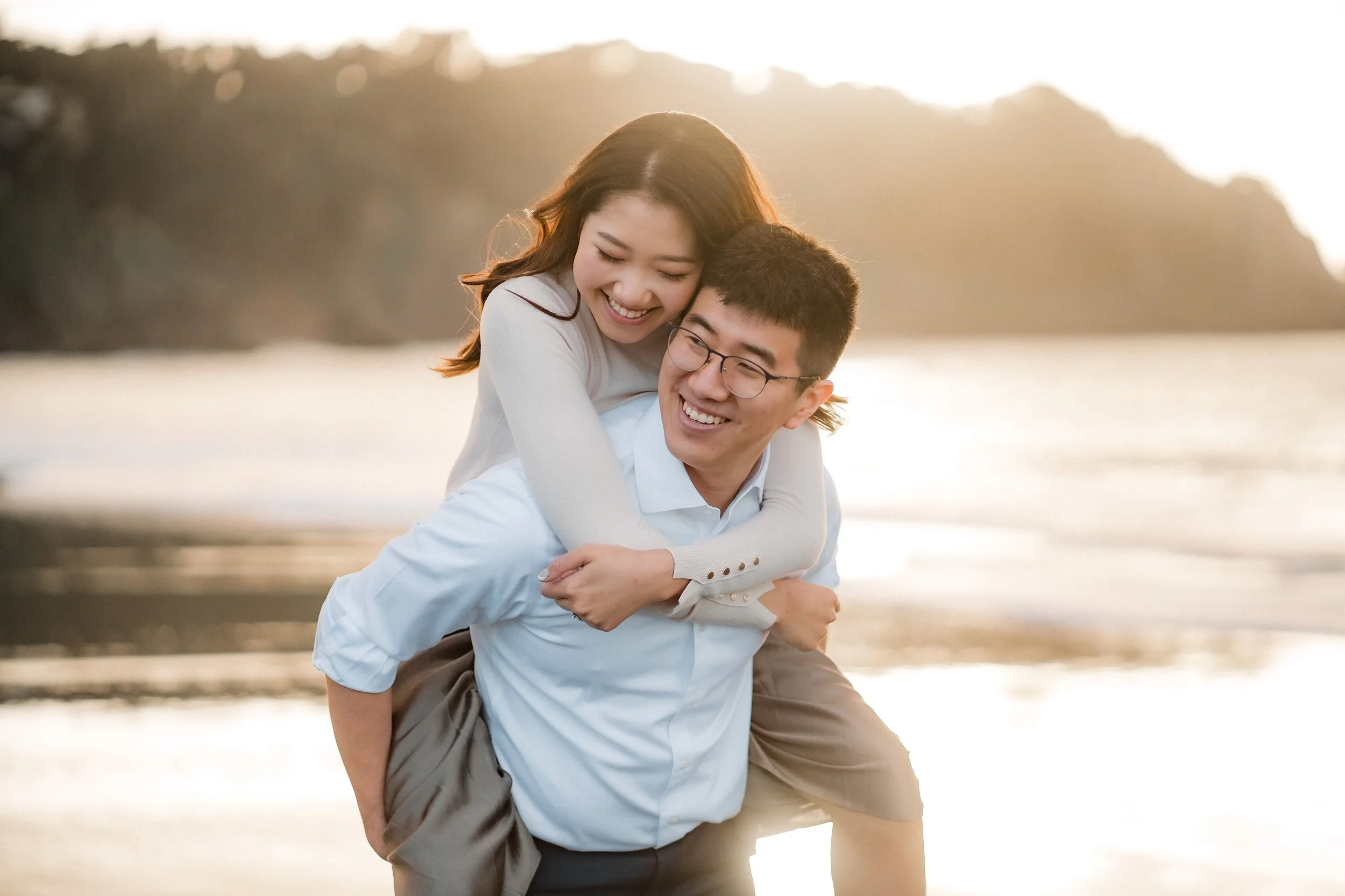 A happy couple at the beach during sunset, with the woman on the man's back, both smiling and embracing.