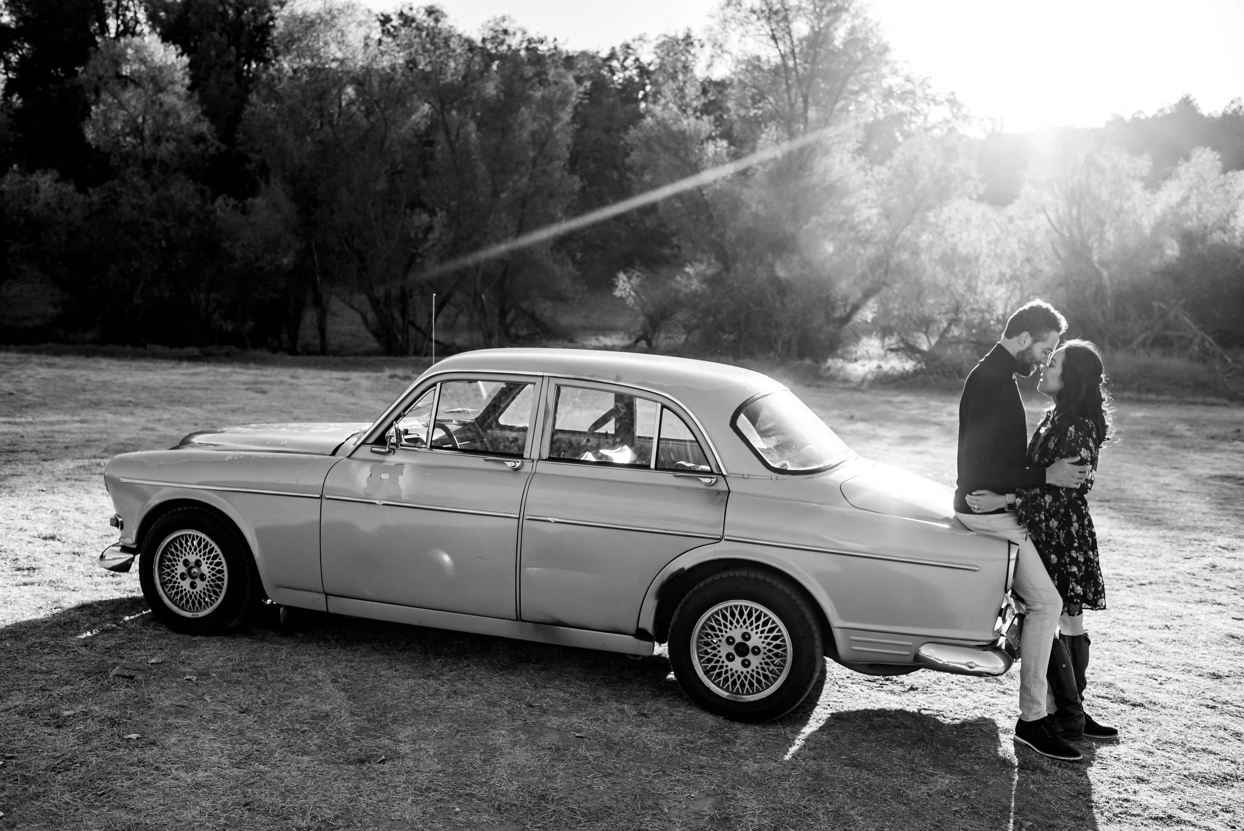 A black and white photo of a couple standing close together in front of a vintage car, outdoors with trees and sunlight in the background, during their engagement session. Photography by Katherine Nicole Photography.