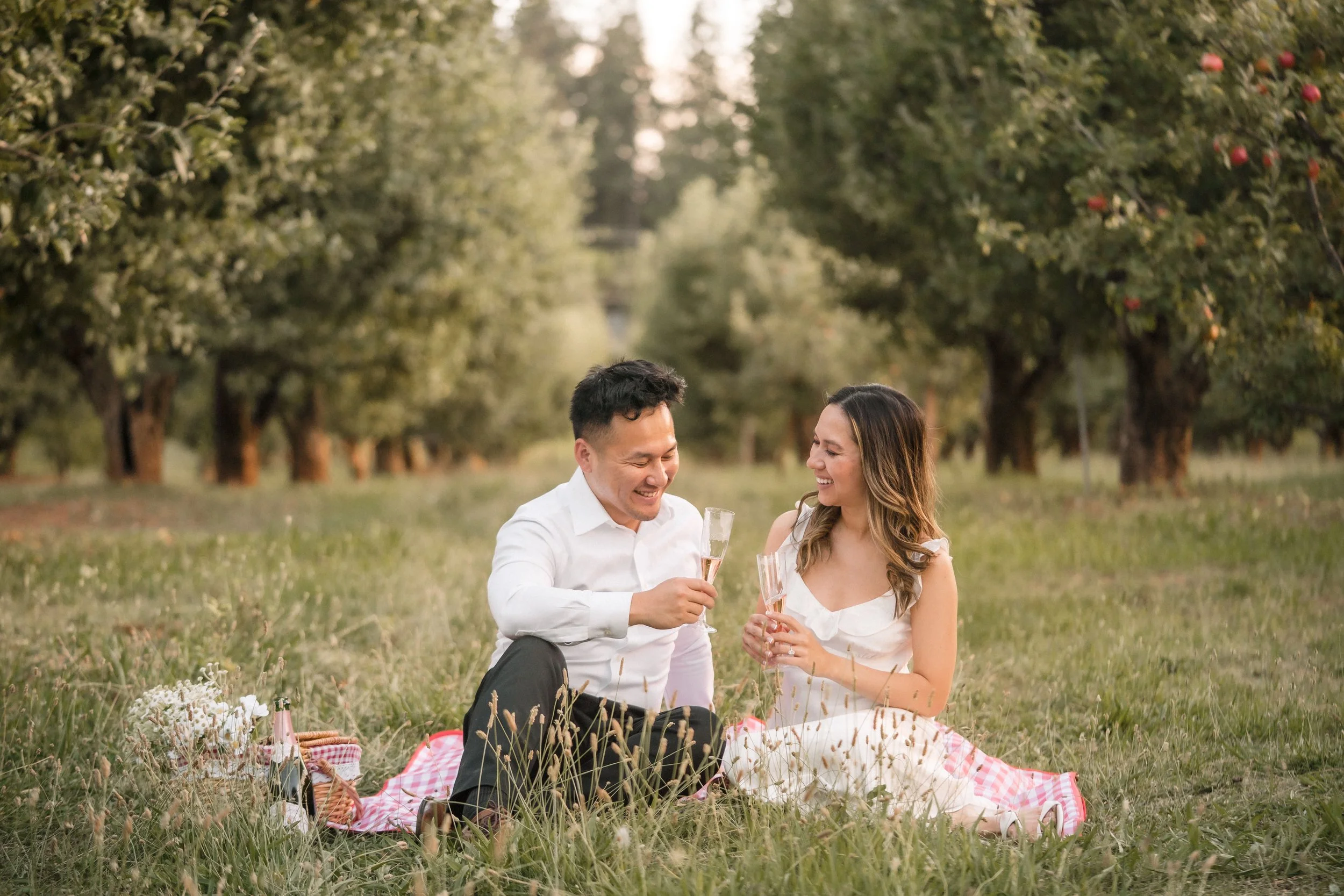 A couple having a picnic outdoors in an orchard, sitting on a blanket and holding champagne glasses, smiling at each other.