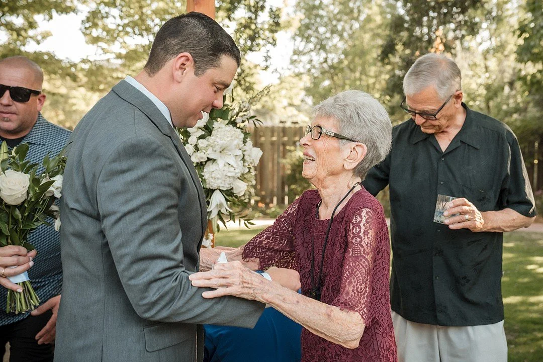 A groom in a gray suit smiling and holding hands with an elderly woman in a burgundy lace dress, both smiling at each other during a wedding at the Flower Farm in Loomis, CA. Photography by Sacramento wedding photographer Katherine Nicole Photography
