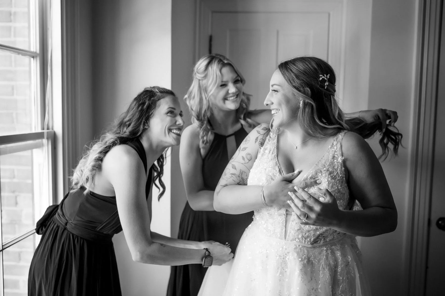 Three women, one in a wedding dress and two in formal dresses, smiling and hugging each other near a window at the Monte Verde Inn. Photo by Sacramento wedding photographer Katherine Nicole Photography.