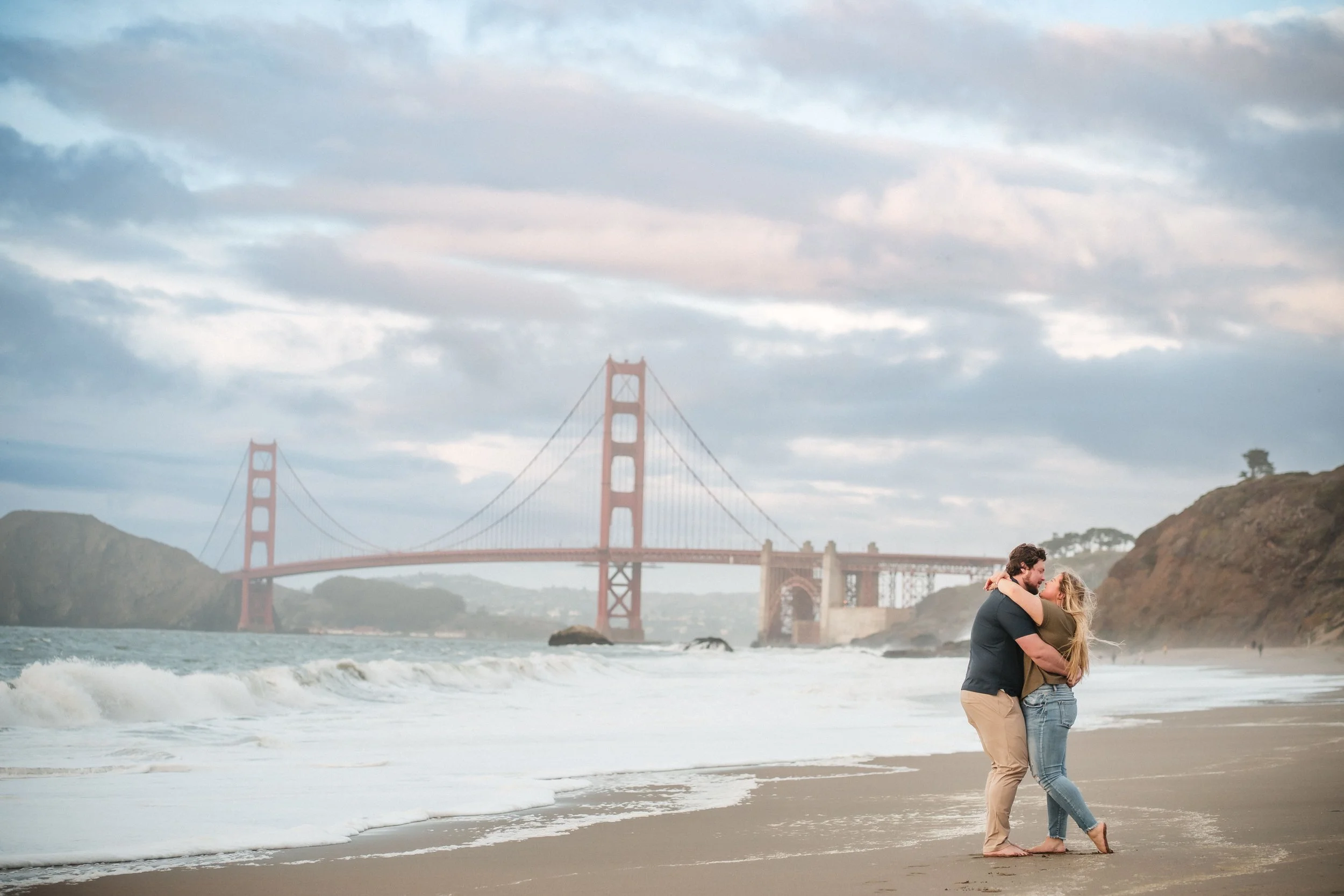 A couple embracing and kissing on a beach with the Golden Gate Bridge in the background under a cloudy sky during their Bay Area engagement photos. Photography by Katherine Nicole Photography.