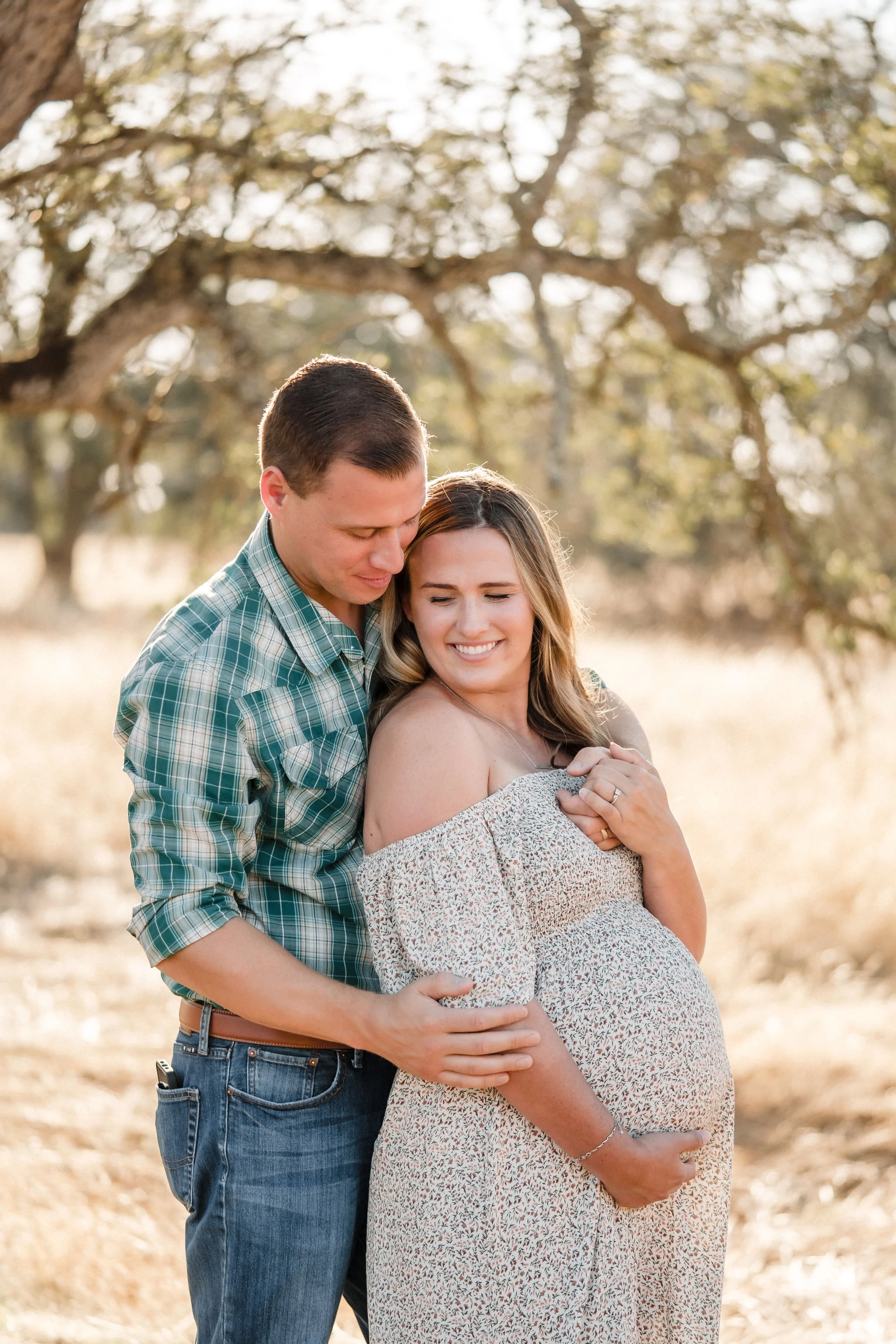 A pregnant woman and a man are standing outdoors with their eyes closed, hugging and smiling, with a tree and sunlight in the background, during a maternity session with Sacramento family photographer Katherine Nicole Photography. 