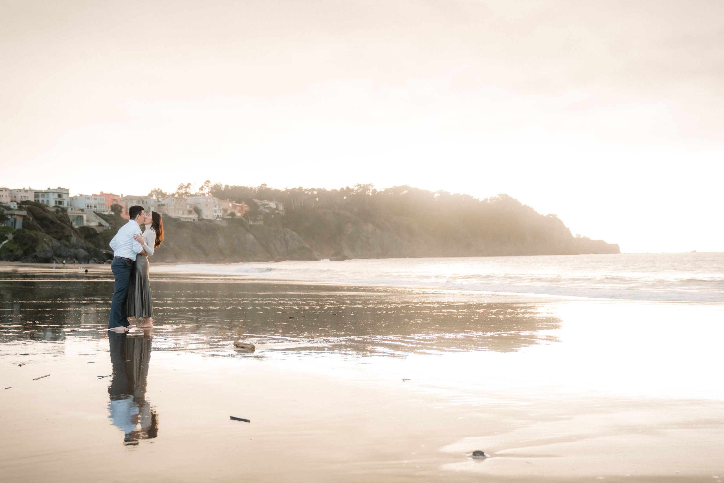 A couple stands close together on a beach during sunset, with their faces nearly touching during their engagement photos at Baker Beach with Katherine Nicole Photography. Their reflection is visible in the wet sand.